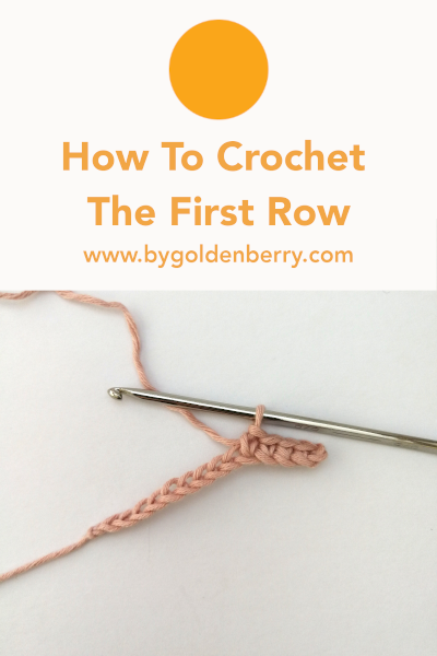 A foundation chain made in pastel pink yarn with a few of the first row of stitches made. The silver crochet hook is still attached to the loop. Above the photo is orange text which reads "How to Crochet the First Row" and "www.bygoldenberry,com".