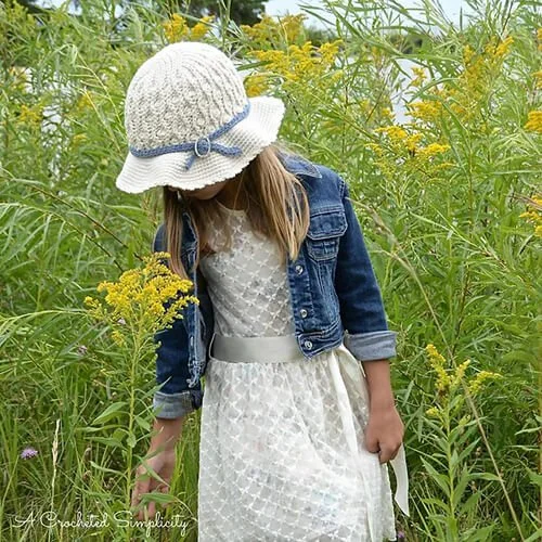 A young girl with shoulder length hair is stood in a field of green. She is wearing a blue denim jacket over a cream coloured dress and the summer saves sunhat. The hat has a distinctive wavy pattern around the body and a blue bandabove the brim.