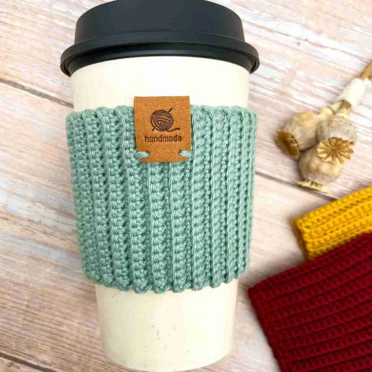 A white coffee cup with a black lid is lying down on a light wooden surface. A light teal, crocheted cup cozy is wrapped around its middle. It has a ribbed pattern and a leather tag stitched onto the middle at the top.