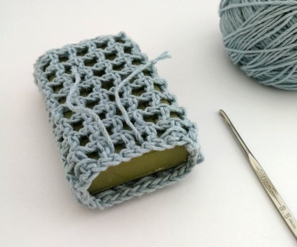The soap saver is sat on a white surface at an angle with the top facing the camera. The top is open and the drawstrings are loose showing the green soap bar on the inside. There is a silver crochet hook on the right hand side.