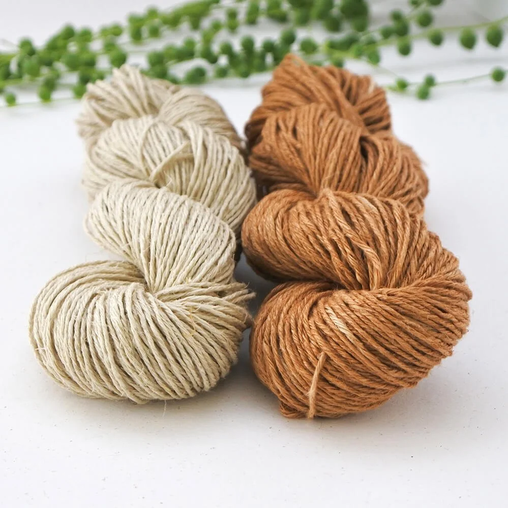 A close up photo of 2 skeins of yarn laying on a white surface. The left is cream and the right is rusty orange. Behind is a green string of pearls plant. The camera is focused on the bottom twists of the skeins.