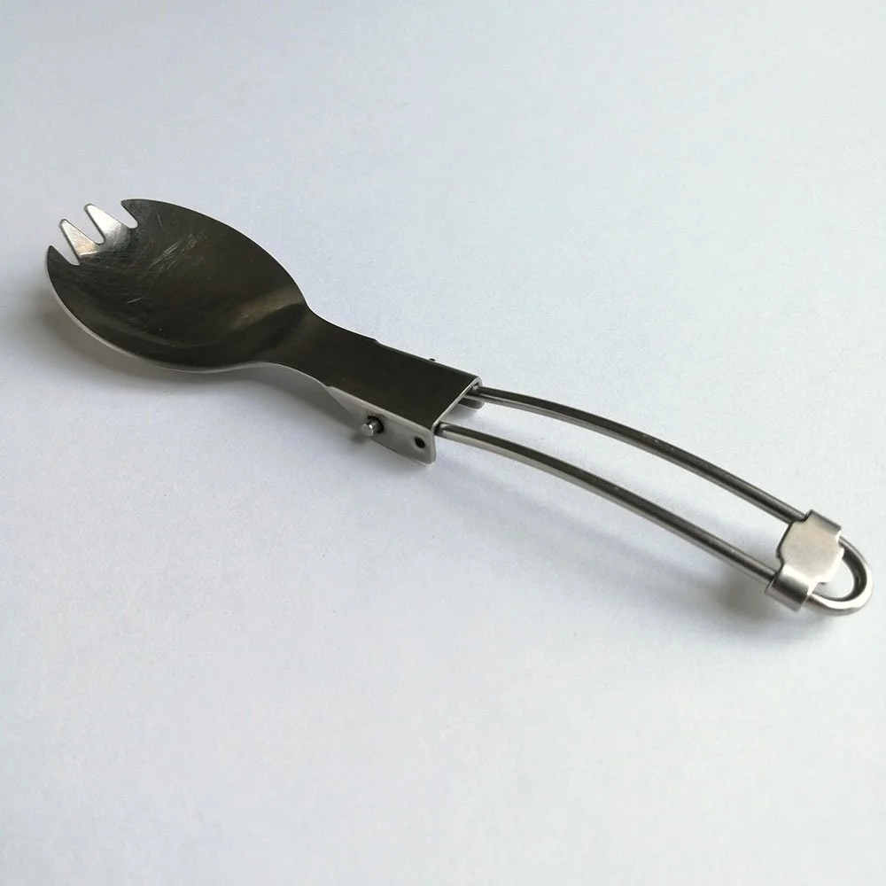 A stainless steel utensil is laying diagonally on a white surface with its top end pointing to the top left corner.  The top end is round like a spoon but had four prongs like a fork.