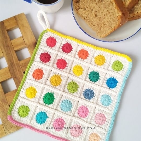 A square pot holder is sat on a white table near a wooden trivet, some bread on a white plate and a small cup of coffee. The pot holder is made up of 5 rows and 5 columns of squares each with a small different coloured round in the center.