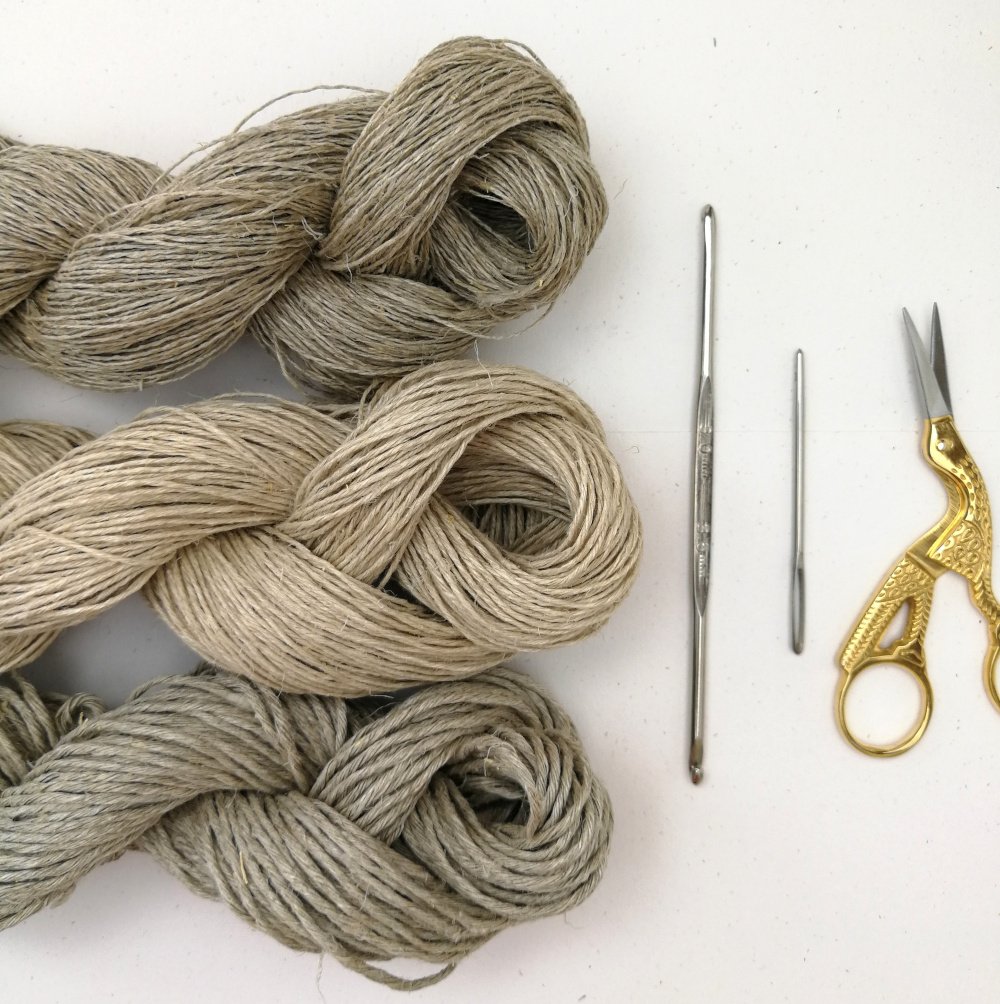 Three skeins of hemp yarn in varying weights are laying horizontally on a white background. They are half into the photo from the left. There is a silver crochet hook, darning needle and a pair of gold and silver stork scissors on the right.