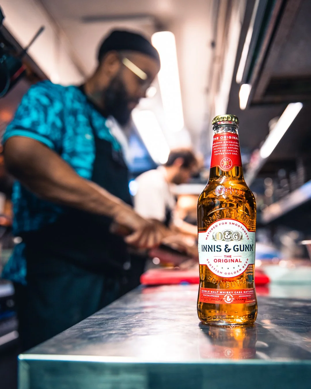 A bottle of Innis & Gunn beer on a metal counter with a blurred chef preparing food in the background.