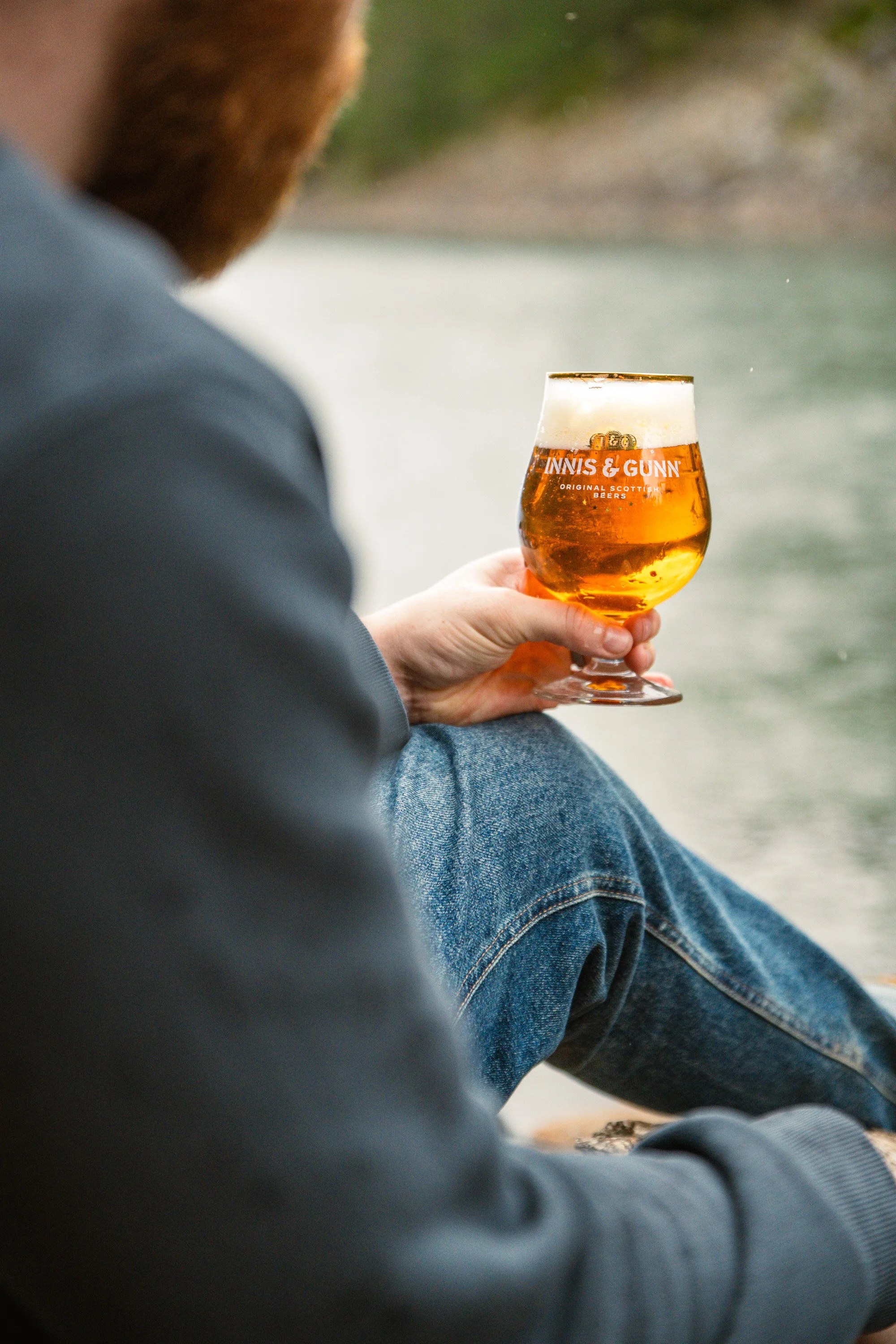 Person sitting by a body of water holding a glass of beer with the label "Innis & Gunn."