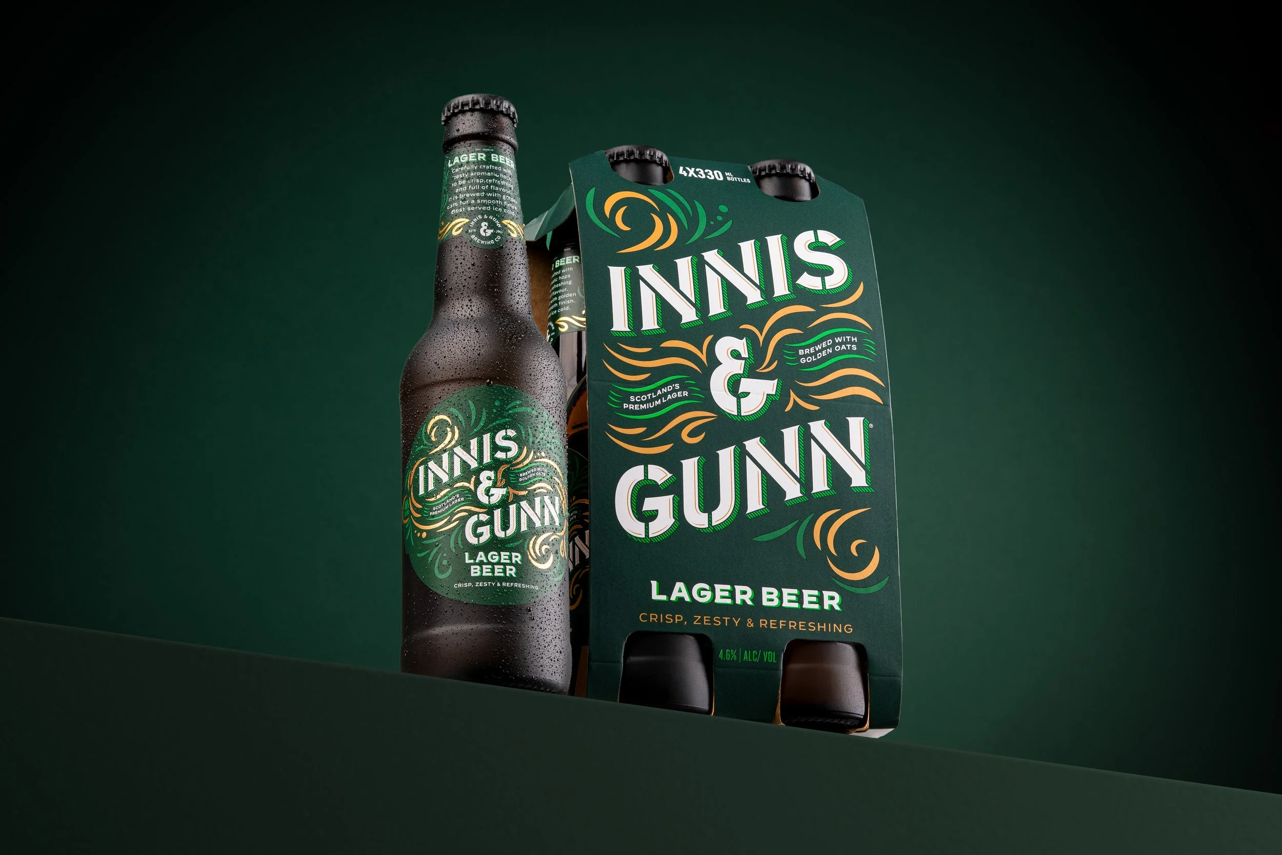 A bottle of INNS & GUNN lager beer and a six-pack carrier of the same brand, with a dark green background and the packaging featuring green, white, and orange design elements.