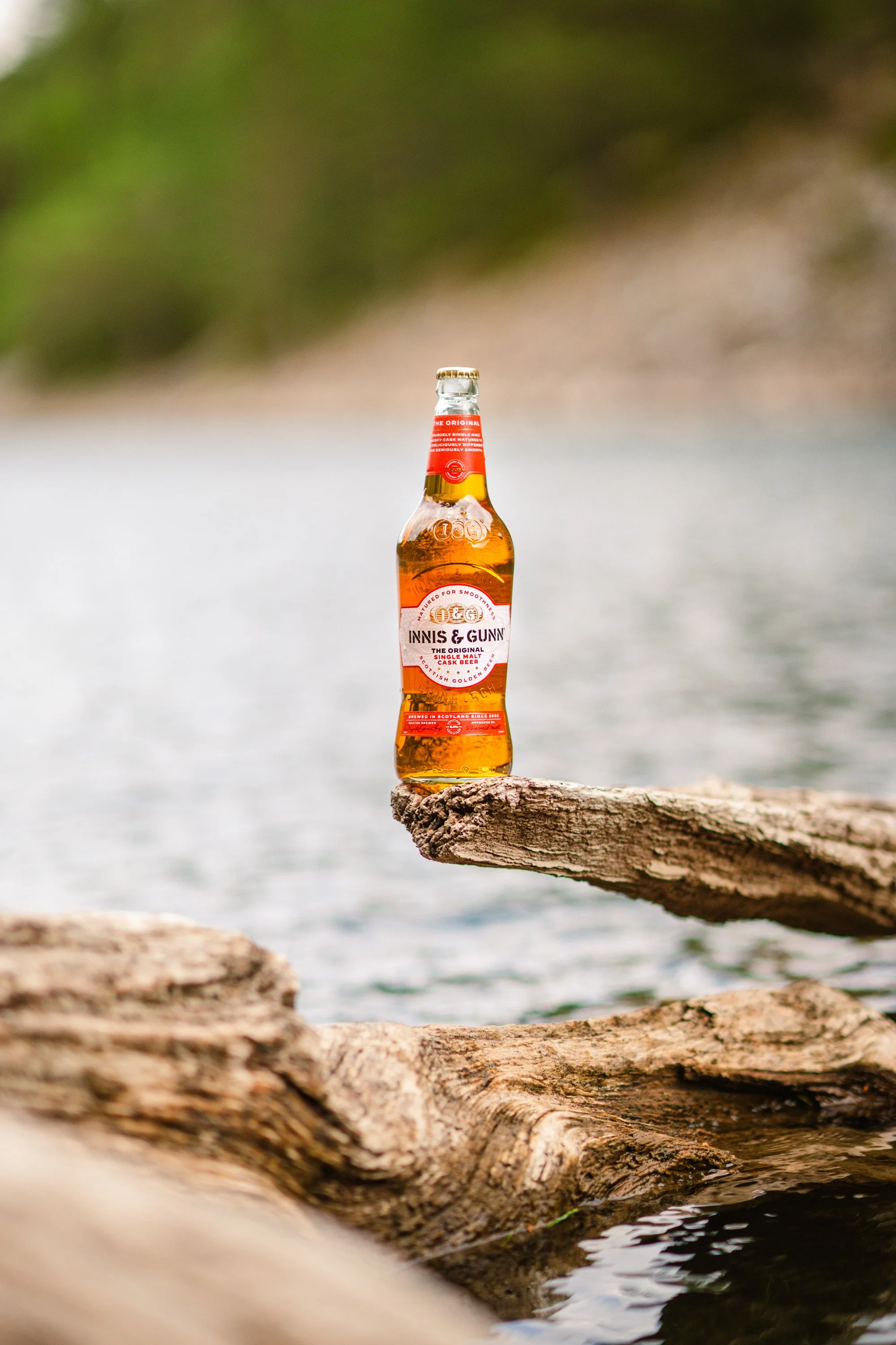 A bottle of Innis & Gunn beer balanced on a piece of driftwood near a body of water with a blurry natural background.