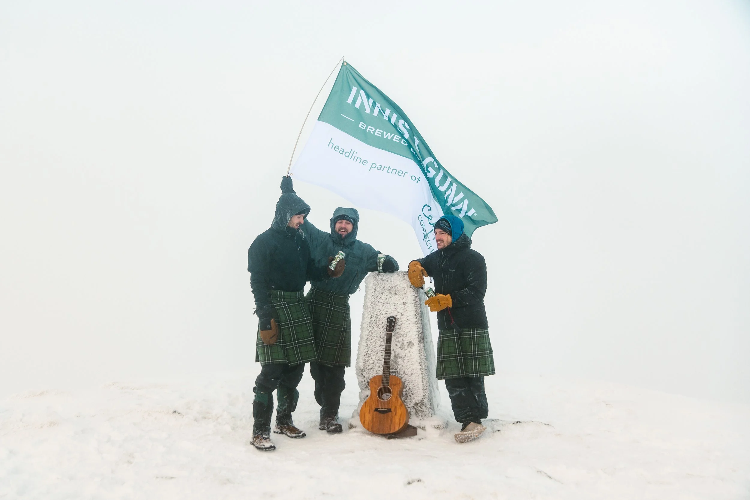 Three men in winter clothing and plaid kilts celebrating on snowy summit with a flag, an ice axe, and an acoustic guitar amidst foggy white background.