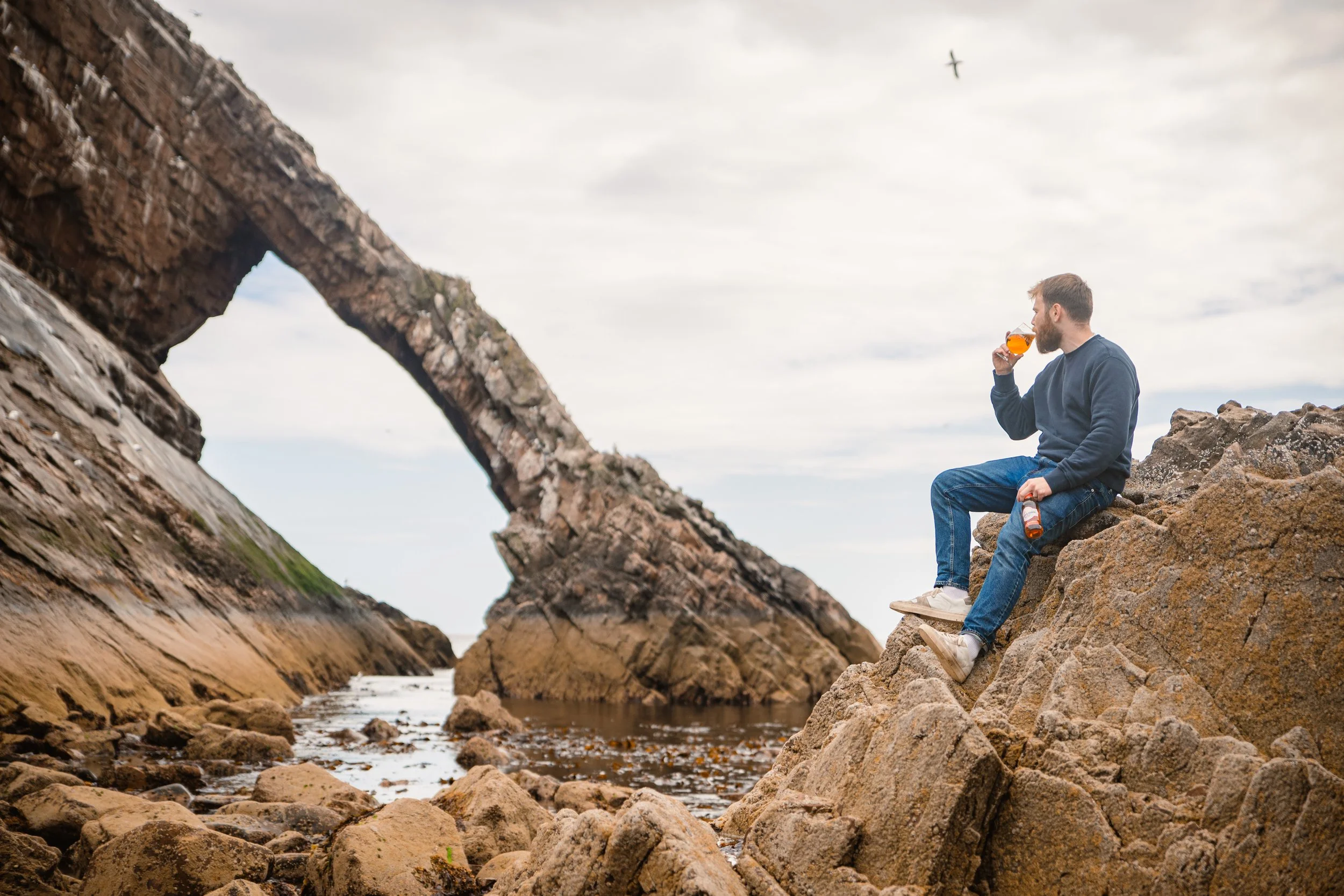A man sitting on rocks near the water drinking a beer at a rocky beach with a natural rock arch in the background.