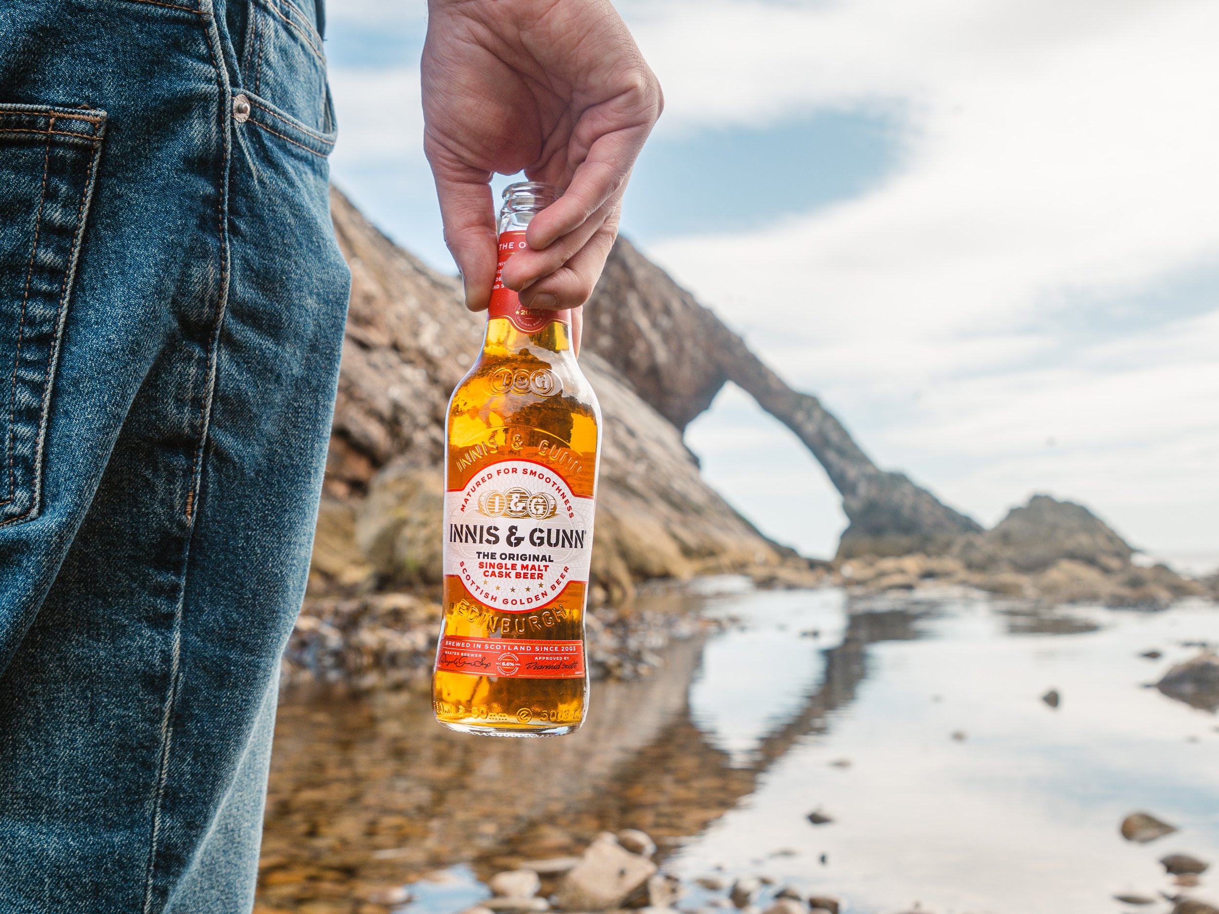 A person wearing blue jeans holding a bottle of Innis & Gunn beer near a rocky beach with a natural rock arch in the background.