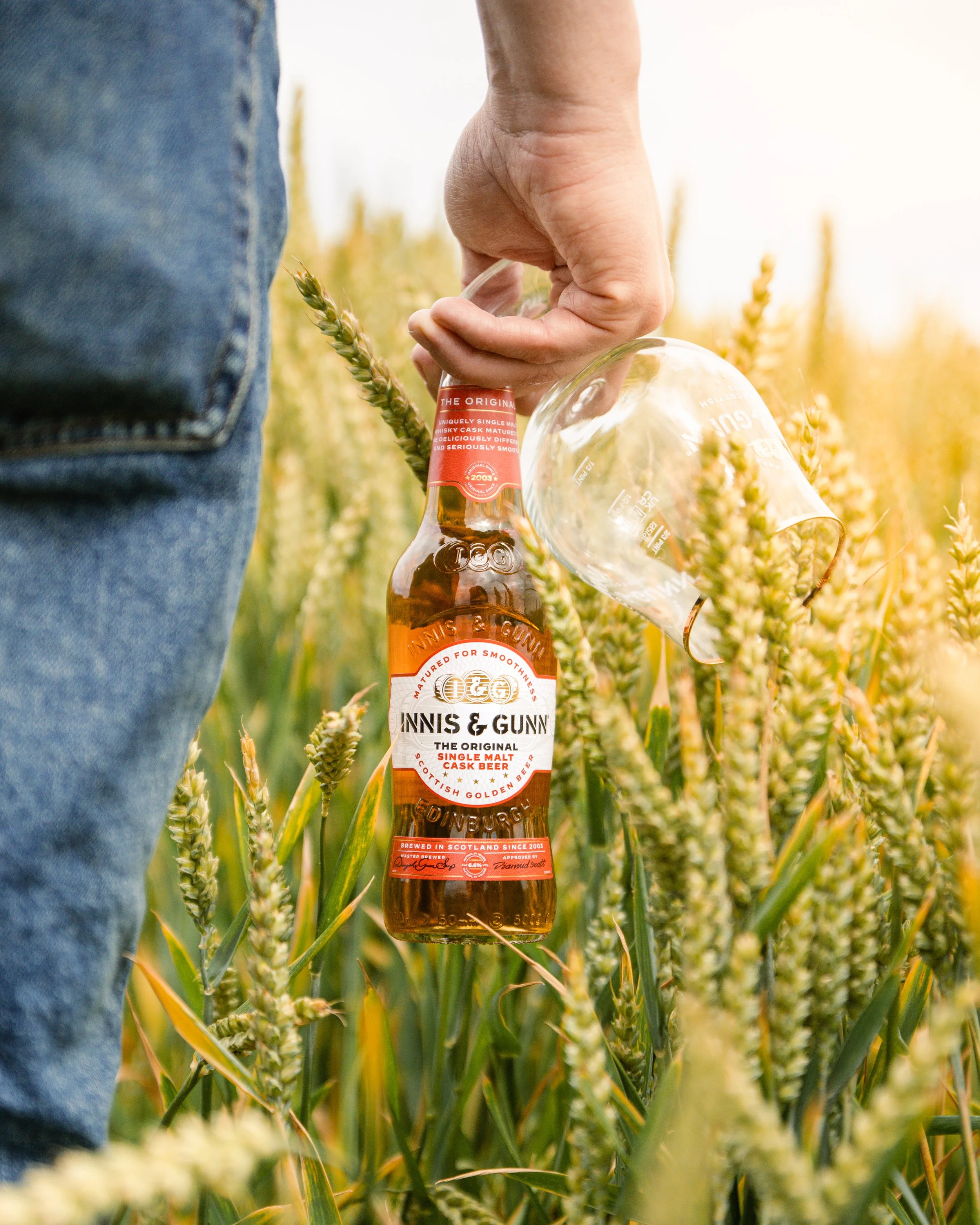A person in jeans holding a bottle of Innis & Gunn beer and a glass in a wheat field.