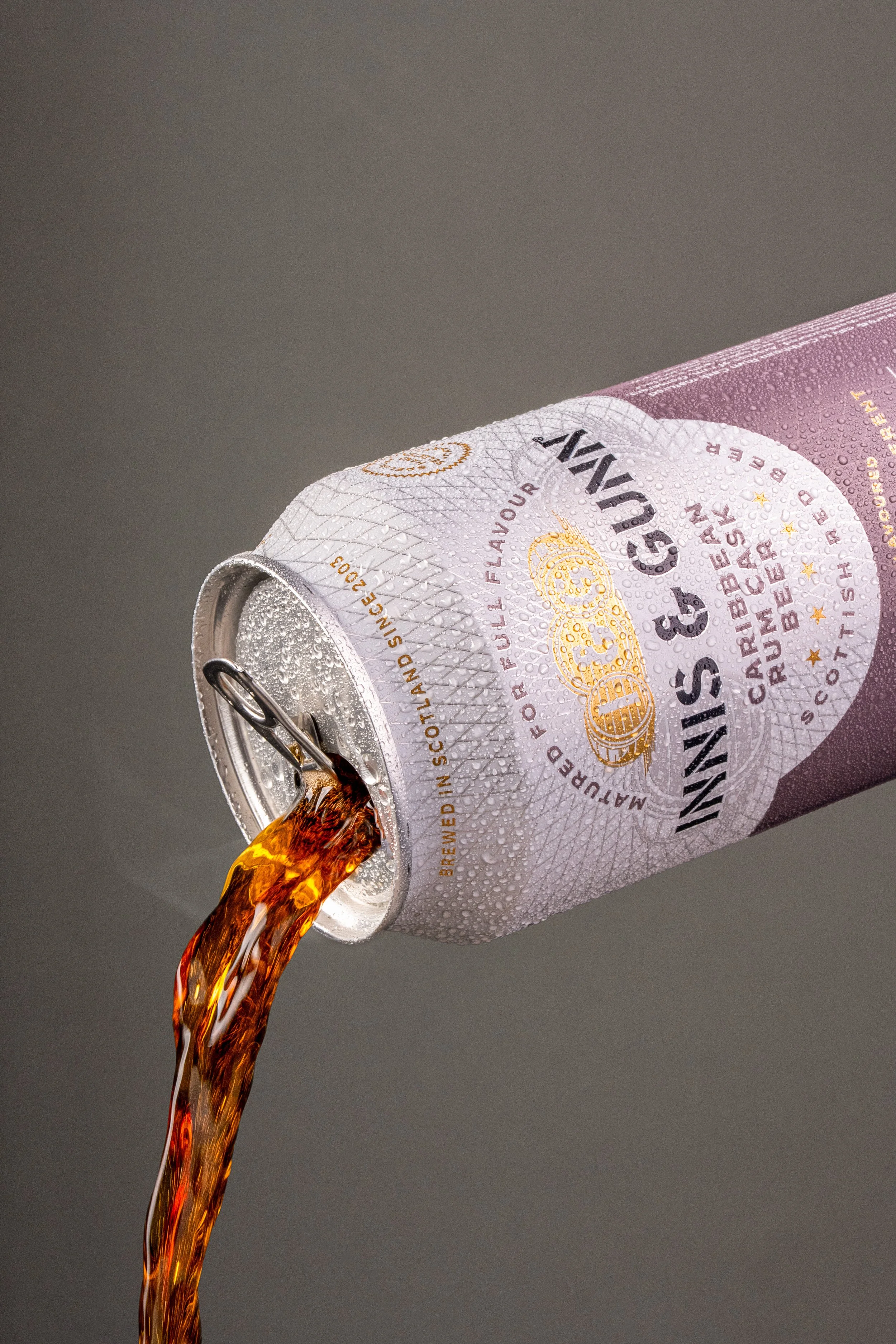 A can of Innis & Gunn beer tilted, pouring out amber-colored beer against a gray background, with droplets of condensation on the can.