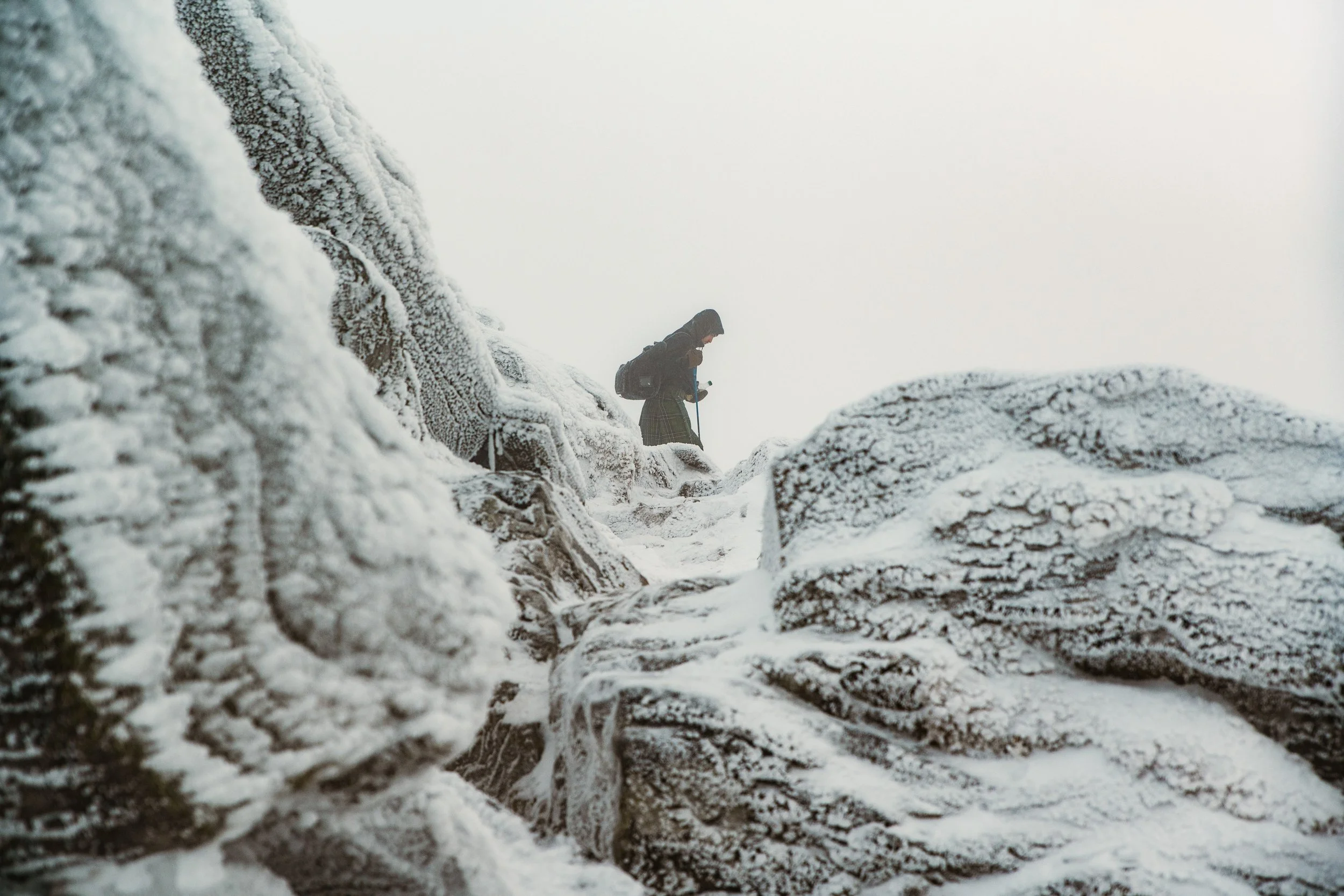 Person hiking on rocky, snow-covered terrain with a snowy, overcast sky.