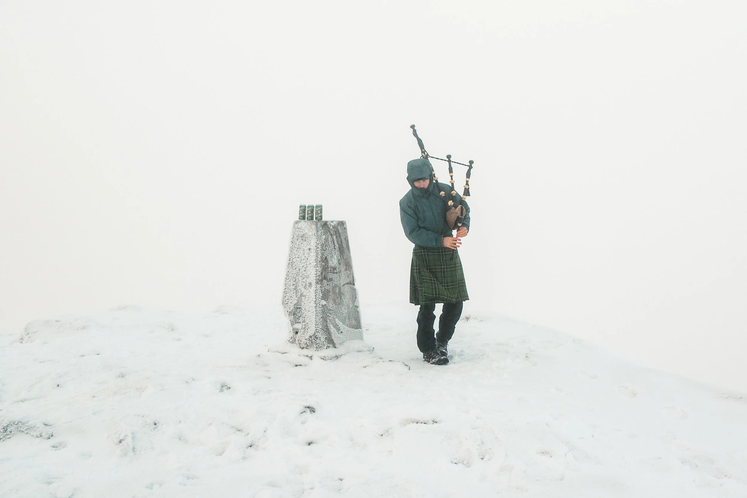 Person in winter clothing playing bagpipes on snowy mountain top with a frost-covered marker and spray painted cans nearby.