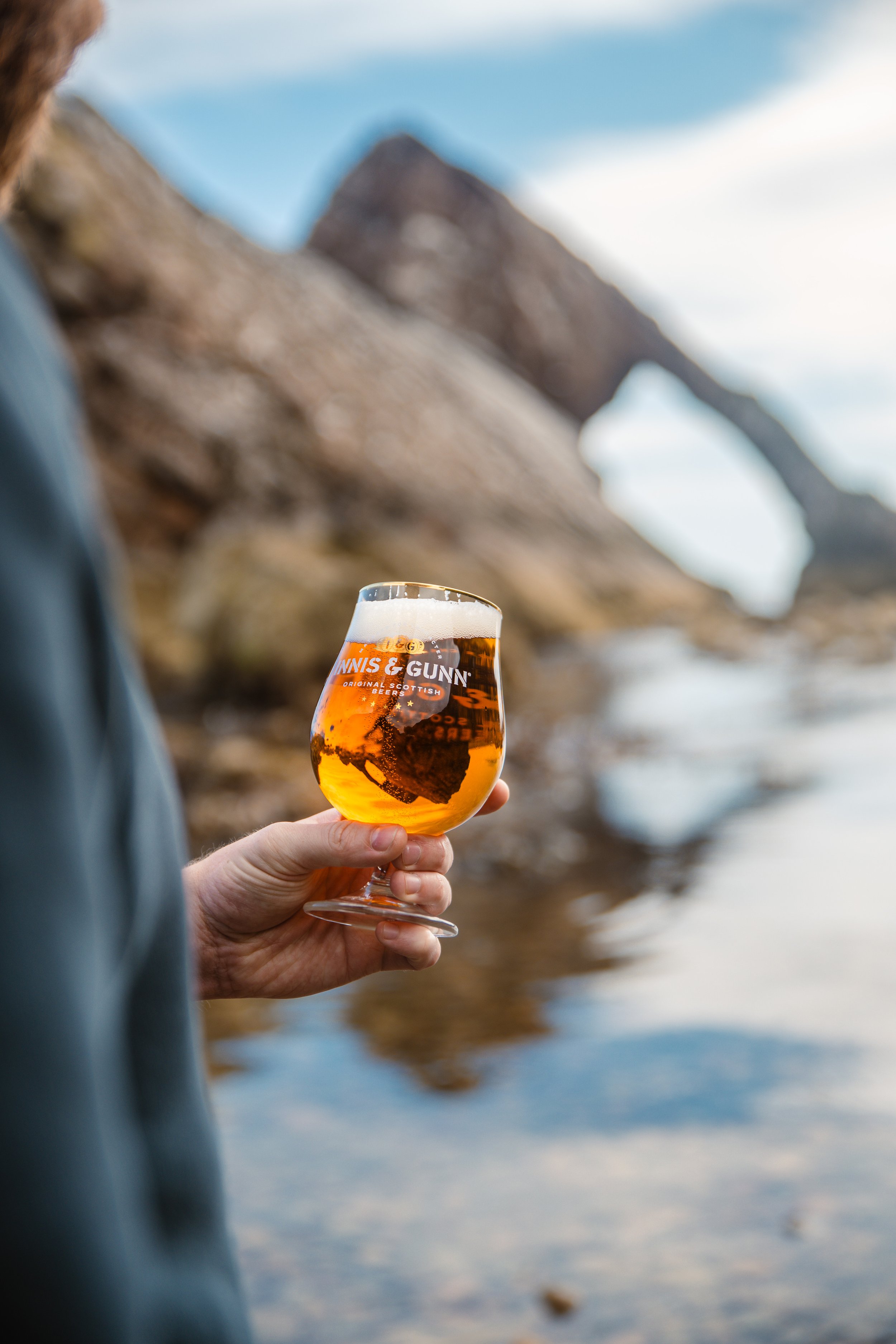 Person holding a glass of amber beer with the logo 'Innis & Gunn' at the beach near rocks and a body of water.