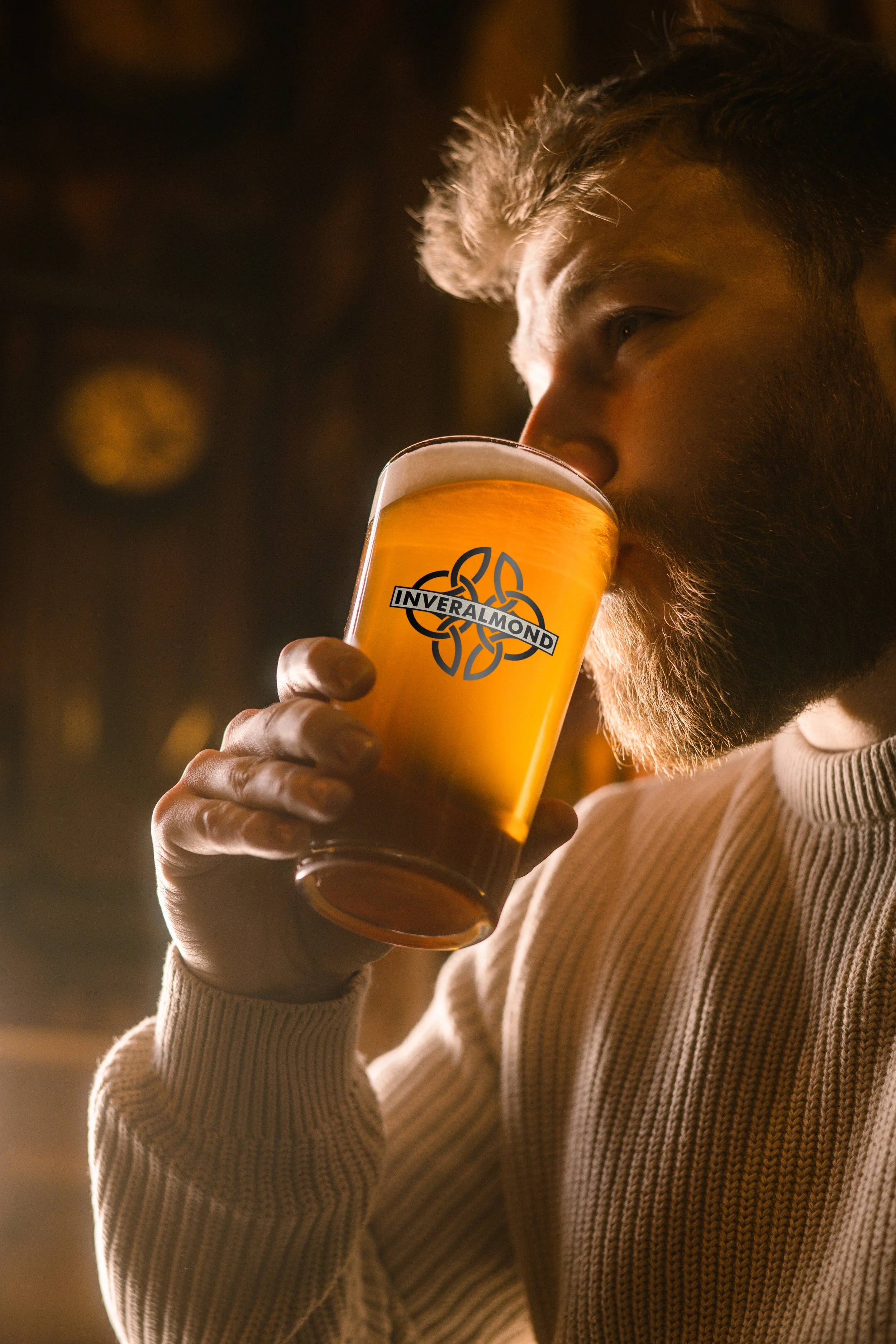 A man with a beard and light-colored hair drinking a pint of beer in a glass with the 'Inveralmond' logo on it, in a dimly lit pub or bar.