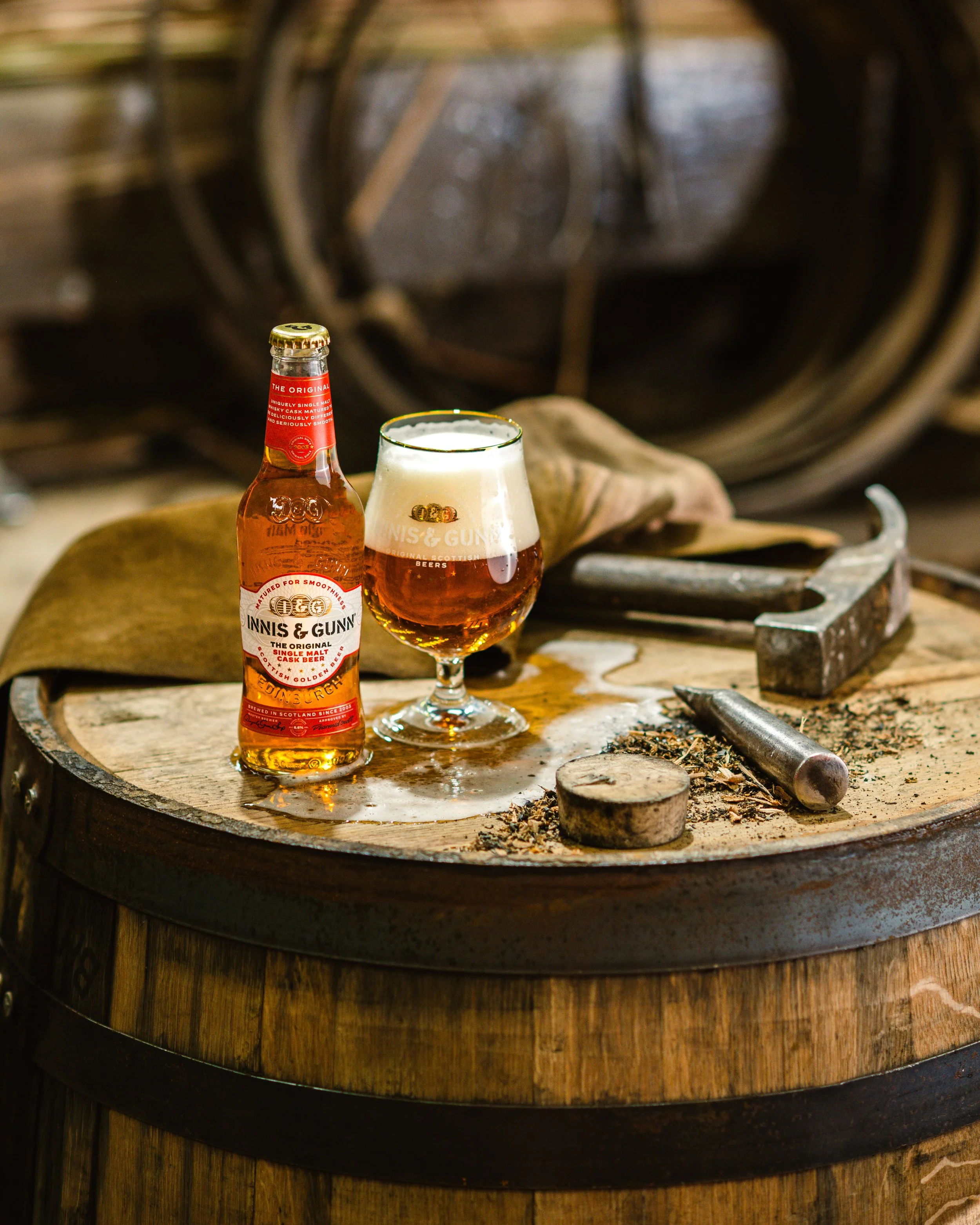 A bottle of Innisc G Gunn single malt scotch whiskey and a glass of amber whiskey with a foamy head on top, placed on a wooden barrel with tools and wood shavings.