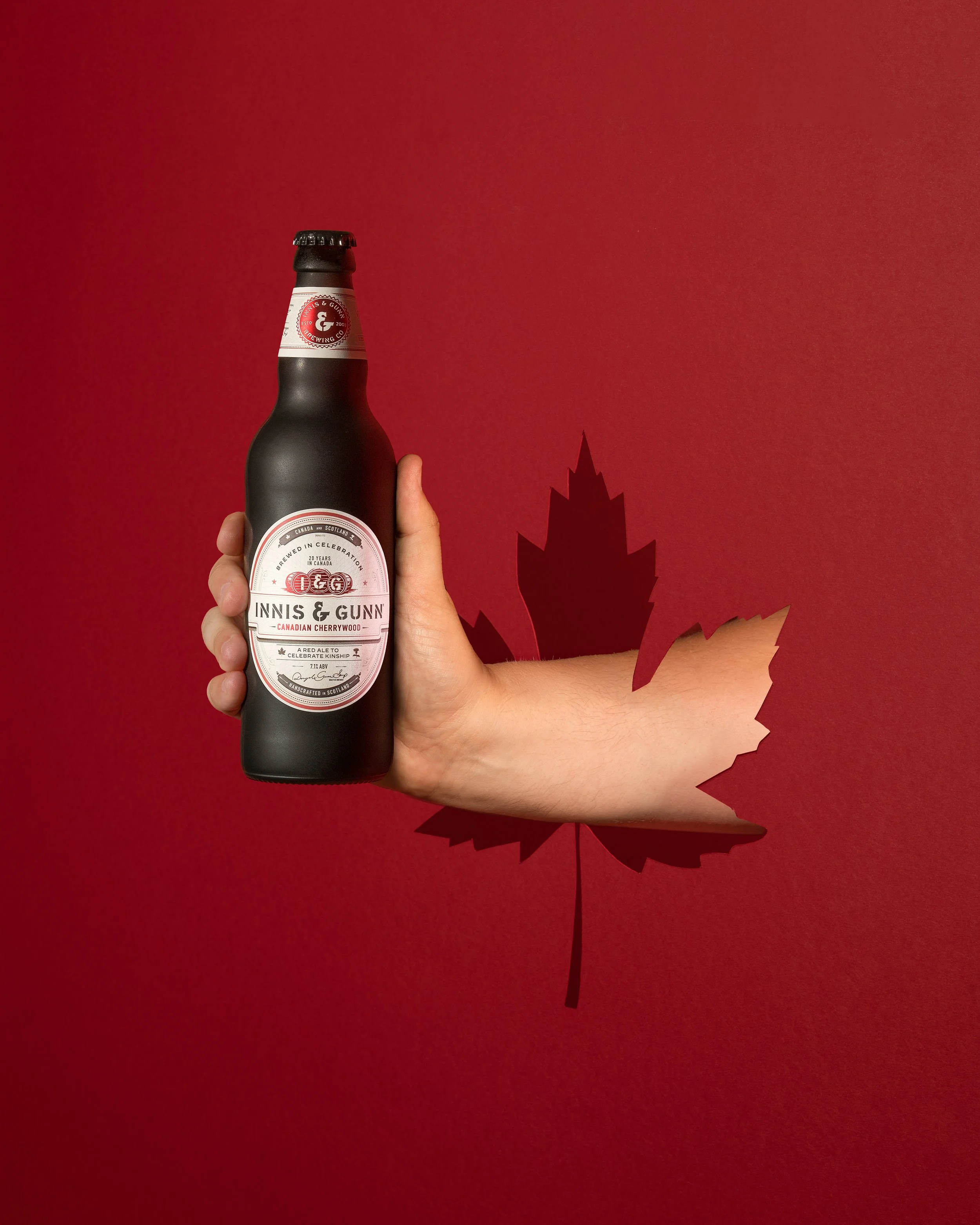 A hand holding a black bottle of Inniskillin & Gunn Canadian Cherrywood beer against a red background with a cutout of a maple leaf.