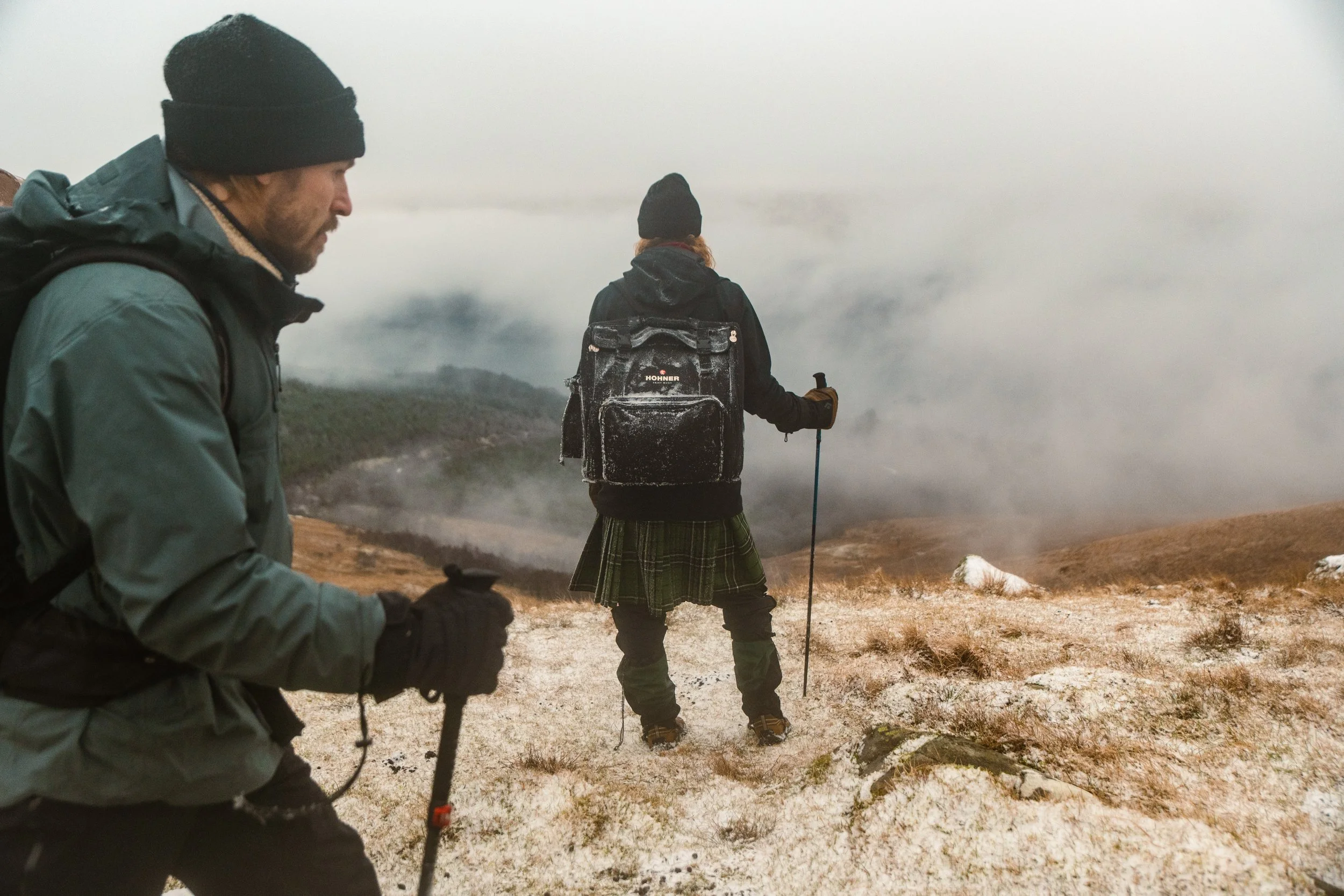 Two hikers wearing winter gear, black beanies, and backpacks standing on a frosty terrain with fog and smoke in the background.