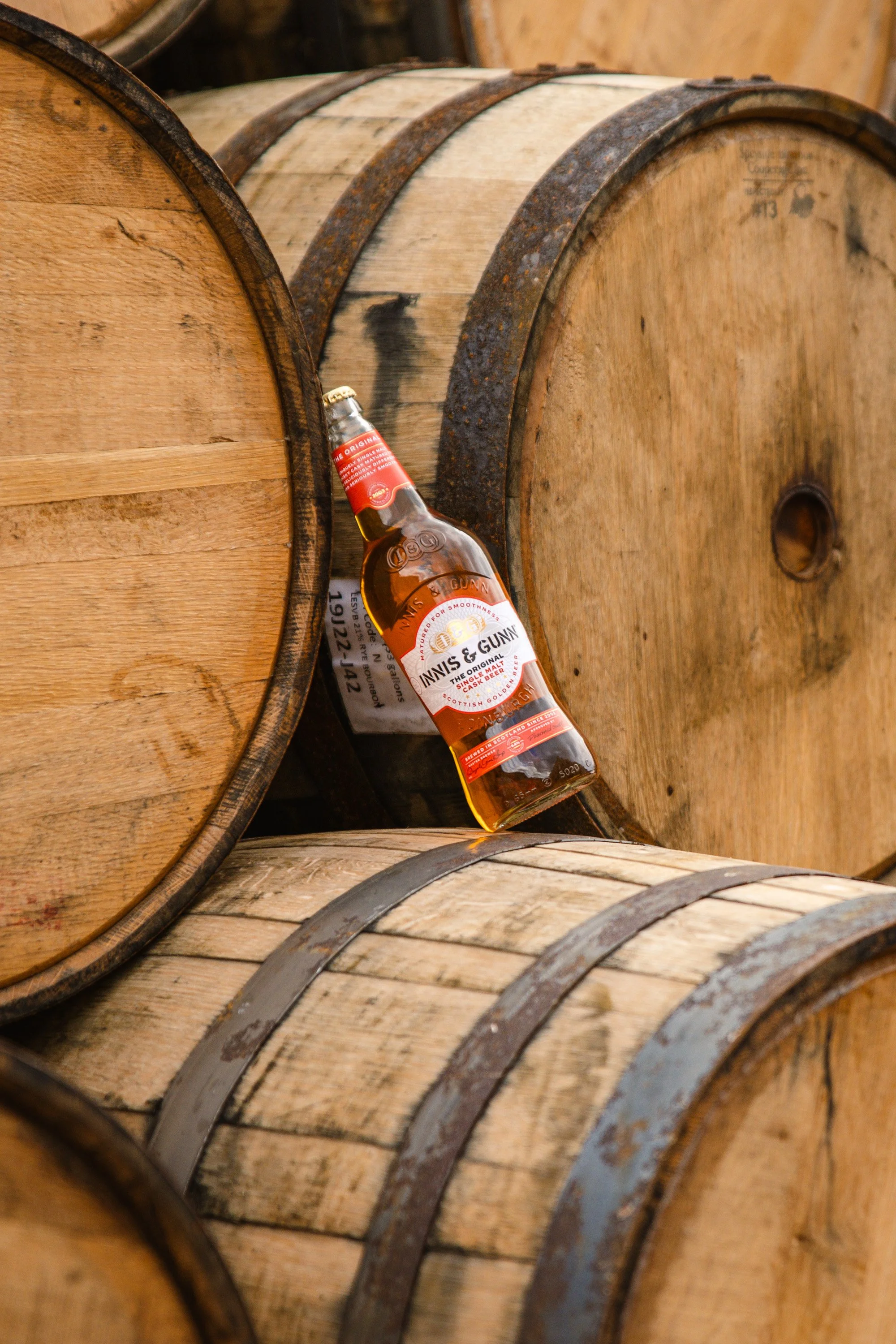 A bottle of Innis & Gunn Scotch Ale leaning against wooden barrels in a cellar or brewery.
