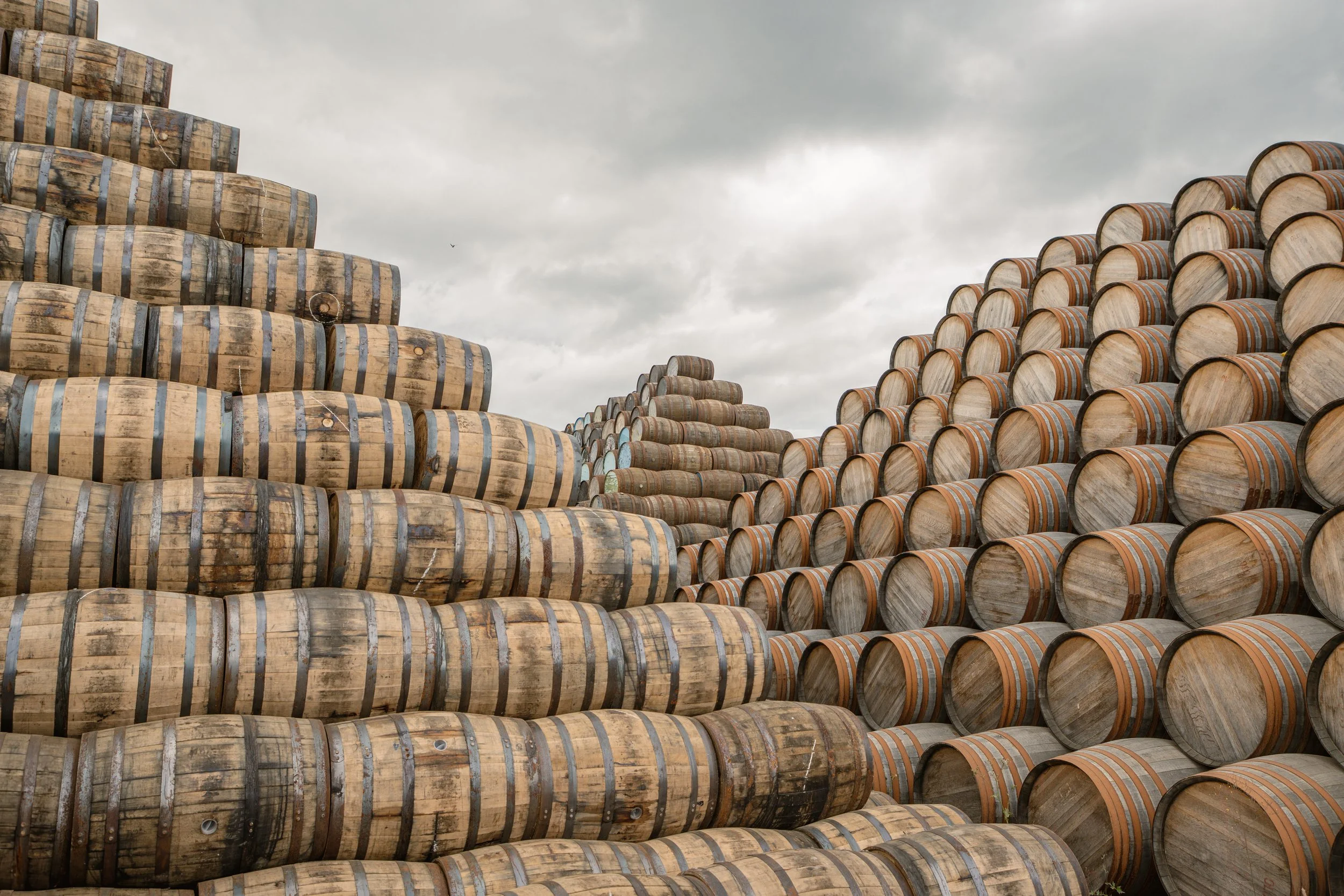 Stacked oak barrels at a distillery or winery against a cloudy sky.