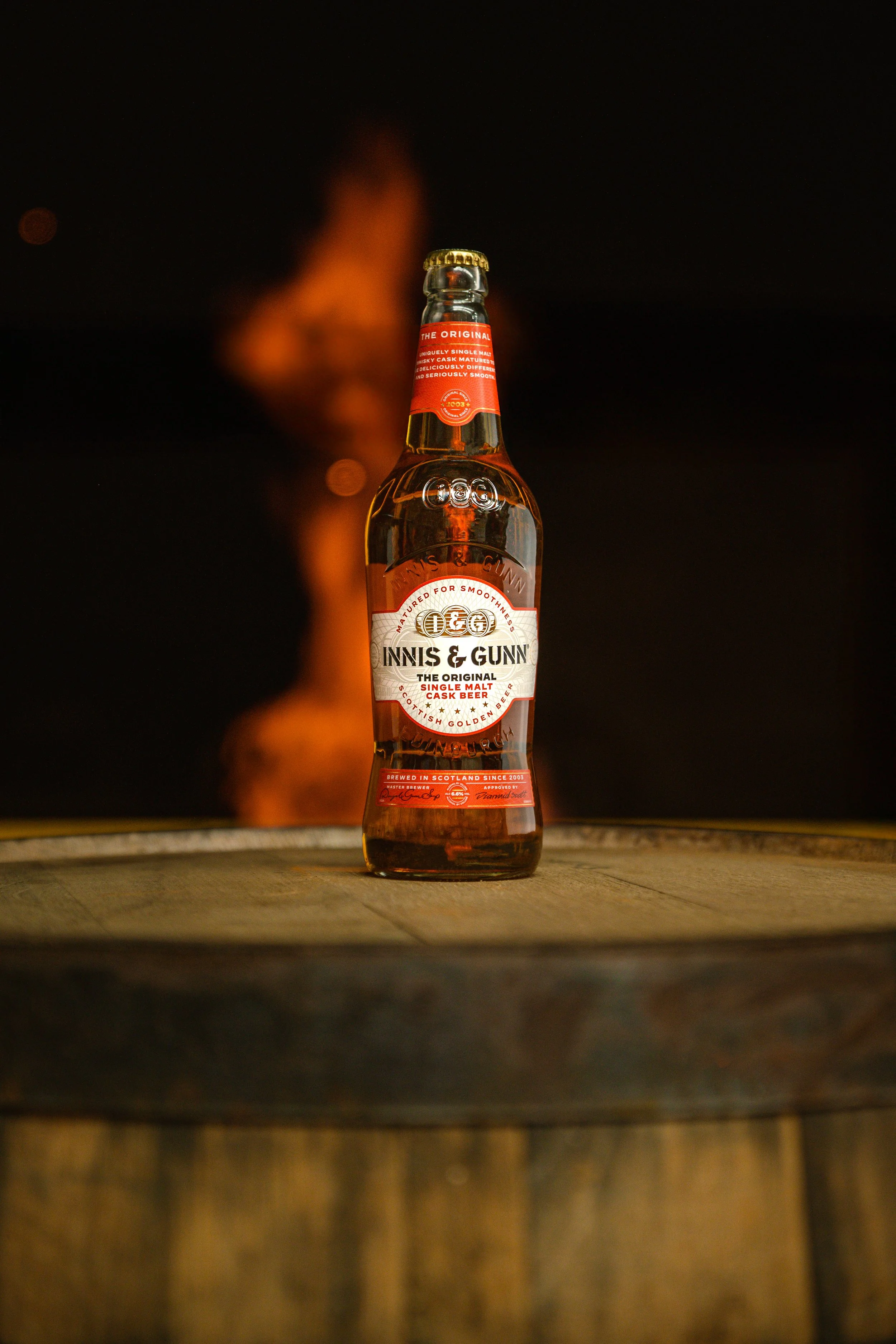 A bottle of Innis & Gunn Scotch Ale on a wooden barrel top with a blurred flame in the background.