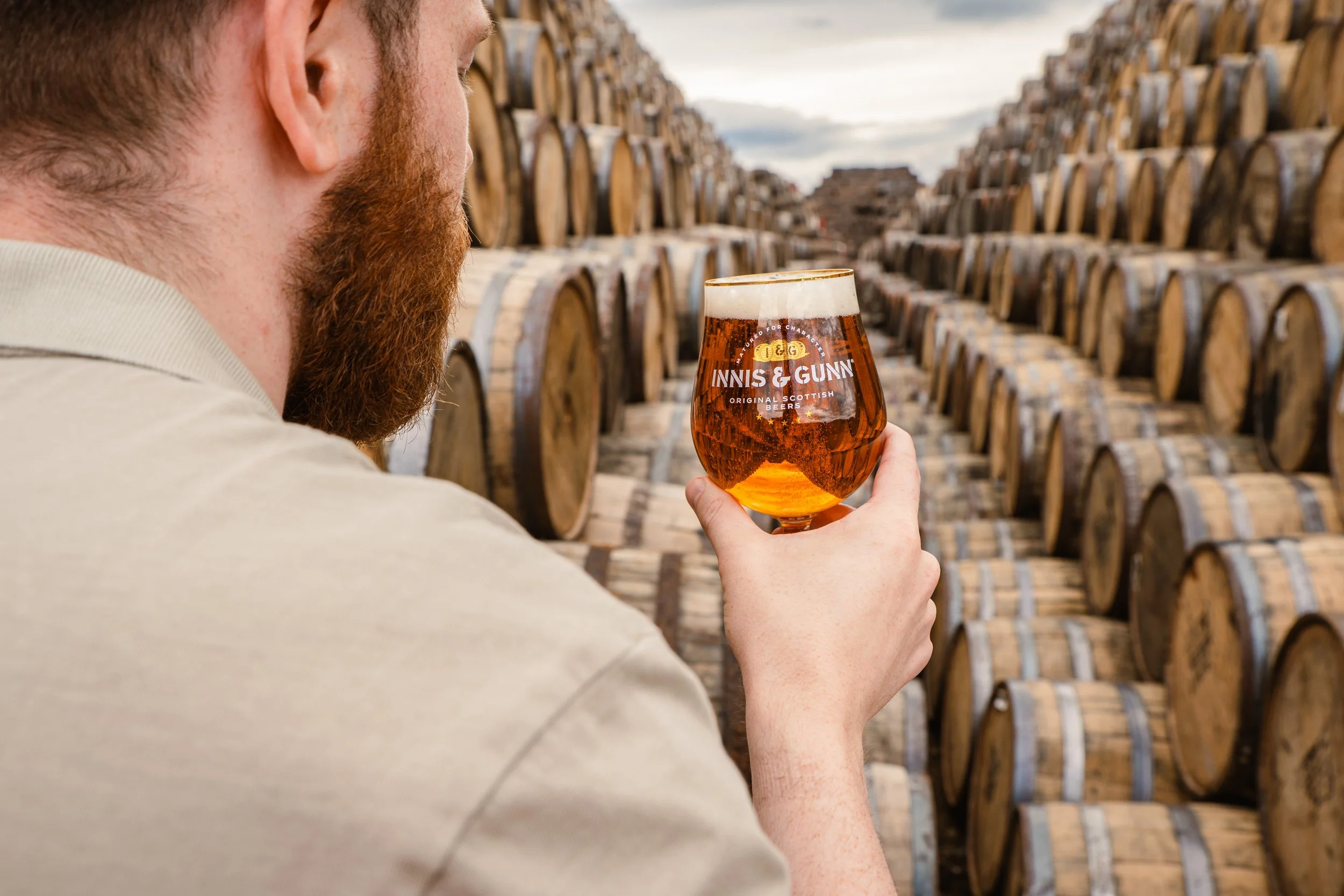 A man with a red beard and light-colored shirt holding a glass of beer in a barrel storage area.