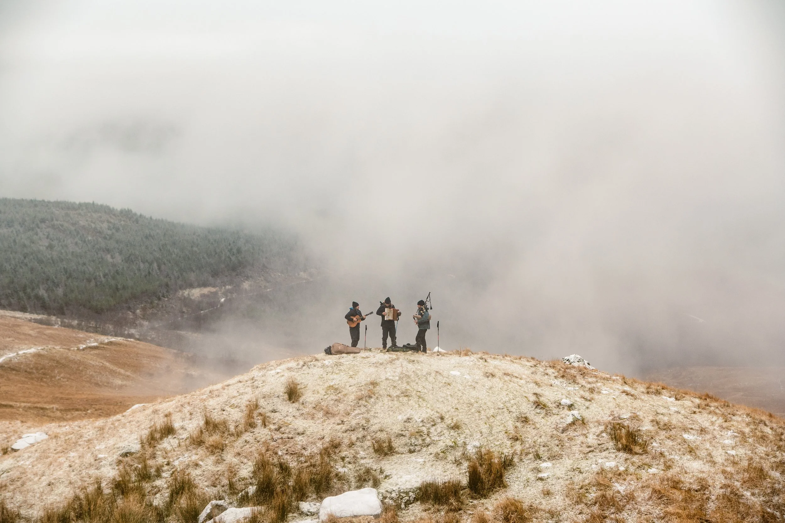 Three musicians performing on a grassy hilltop amid foggy mountains.