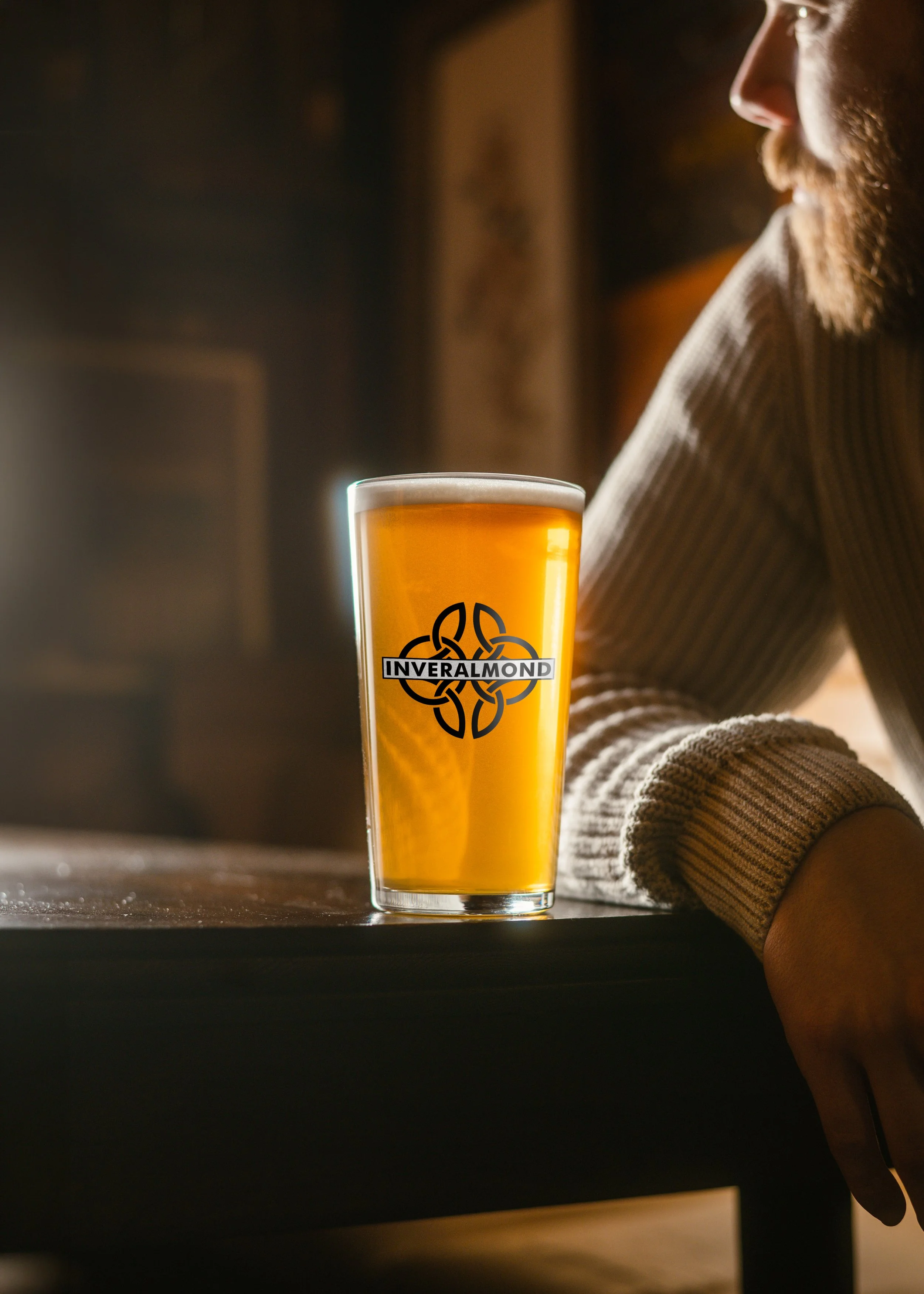 A pint of beer with a logo reading 'Inveralmond' on the glass, sitting on a dark wooden table next to a person wearing a striped sweater, in a warm, softly lit setting.