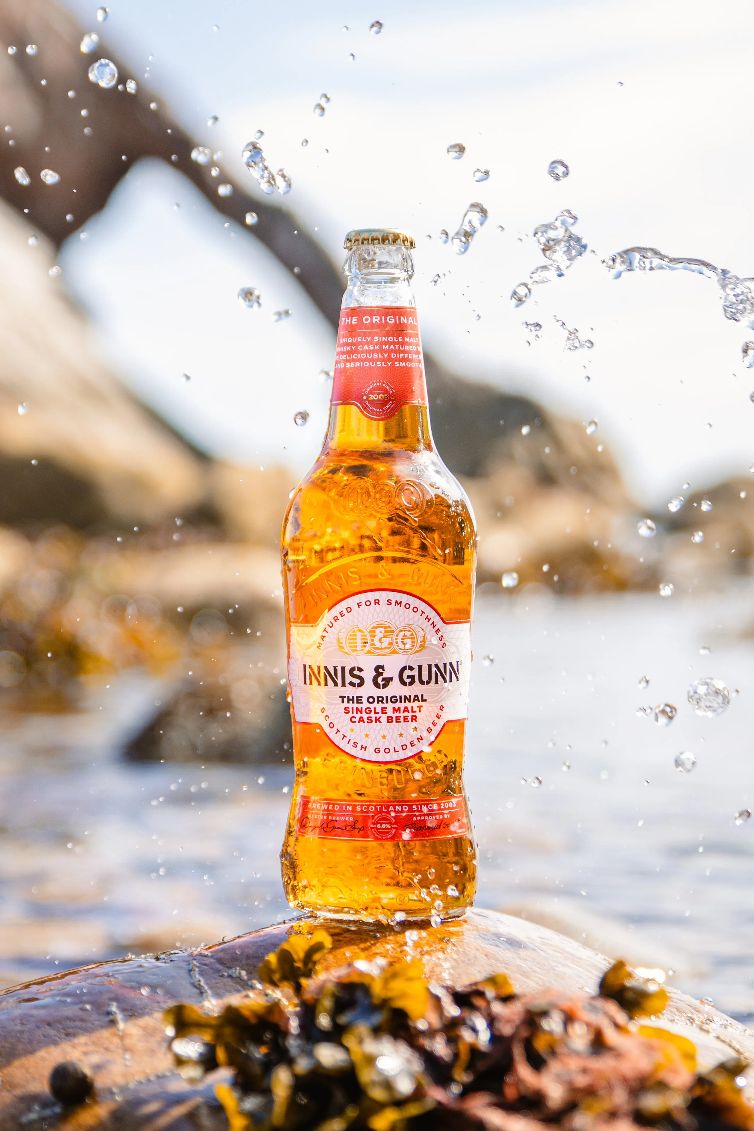 A bottle of Innis & Gunn single malt cask beer standing on a rock near water, with splashes of water around it and a blurred background of rocks.