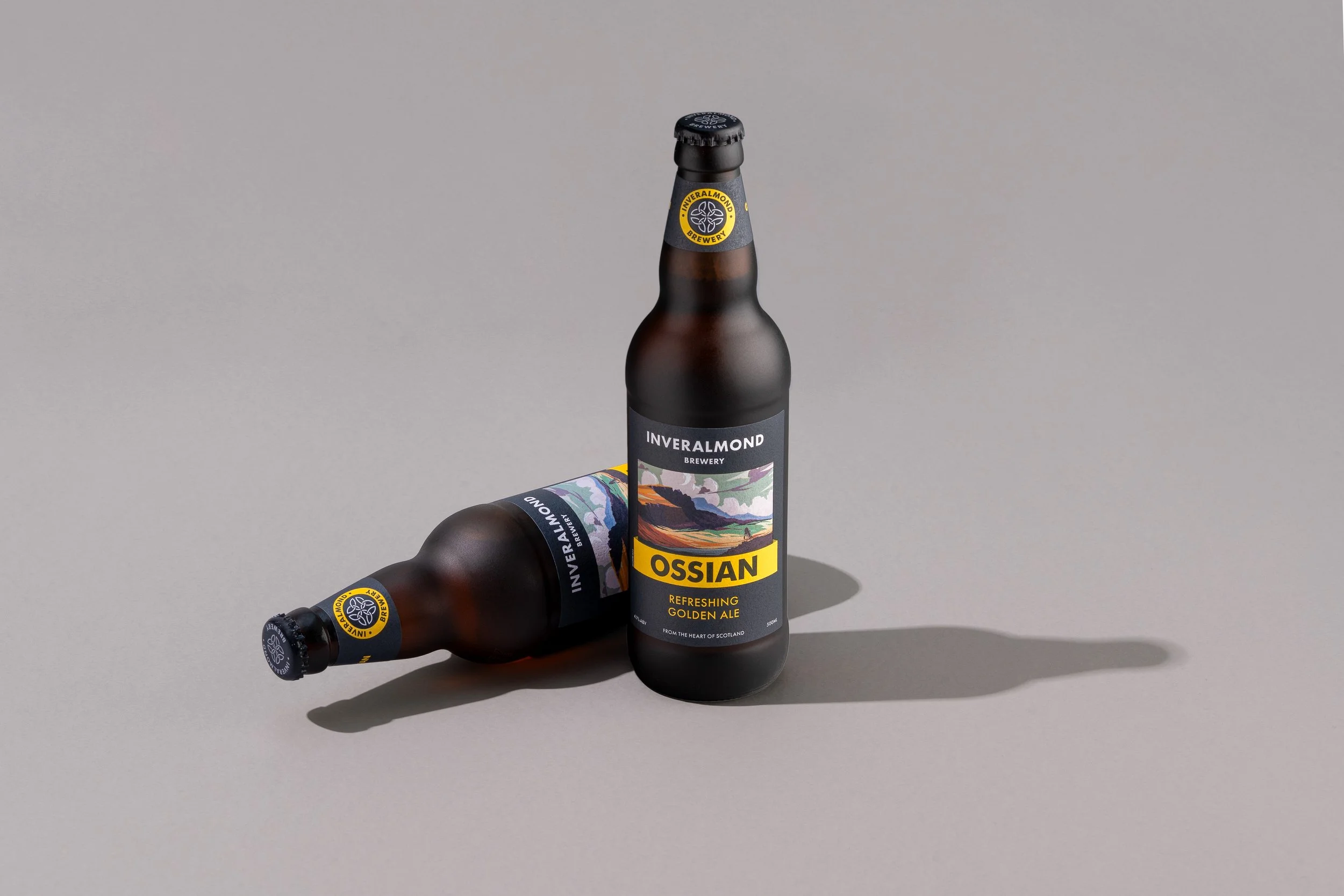 Two bottles of Inveralmond Brewery Ossian beer, one standing and one lying down, on a neutral gray background with shadows.