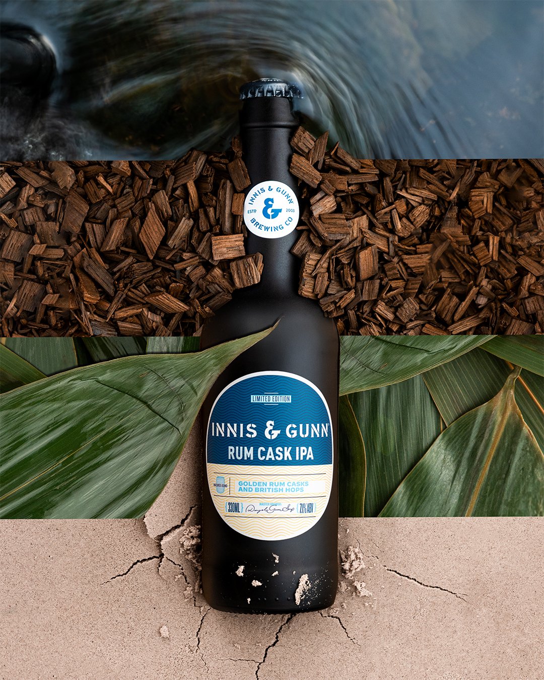 A black bottle of Inniss & Gunn Rum Cask IPA beer lies on sandy ground, surrounded by green leaves, with a bottle cap and label indicating limited edition with gold rum casks and British hops, and the alcohol content of 7.4% ABV.