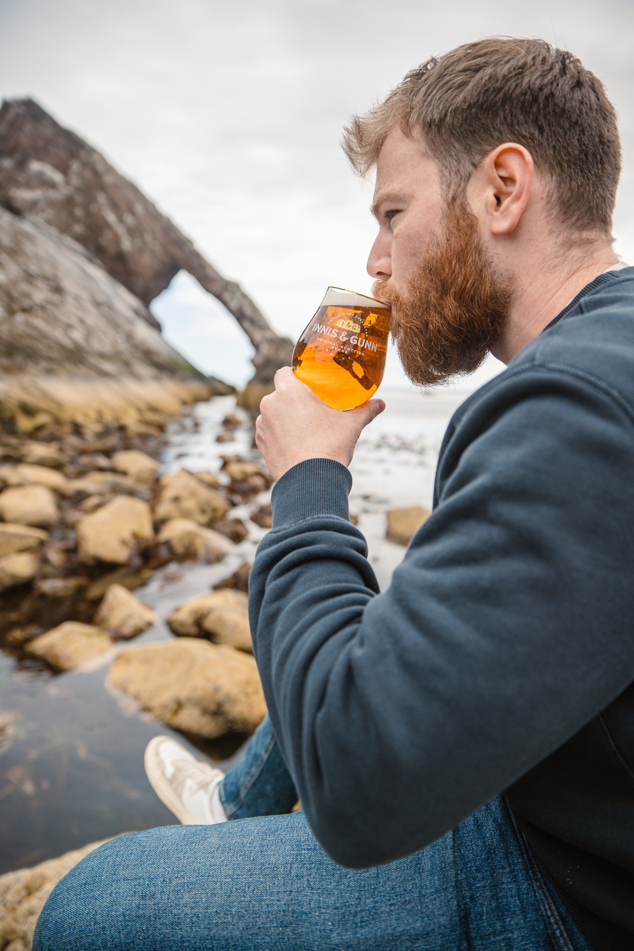 Man with a beard sitting by rocks near a beach, drinking beer from a glass with INNIS & GUNN branding, with a rock arch formation in the background.