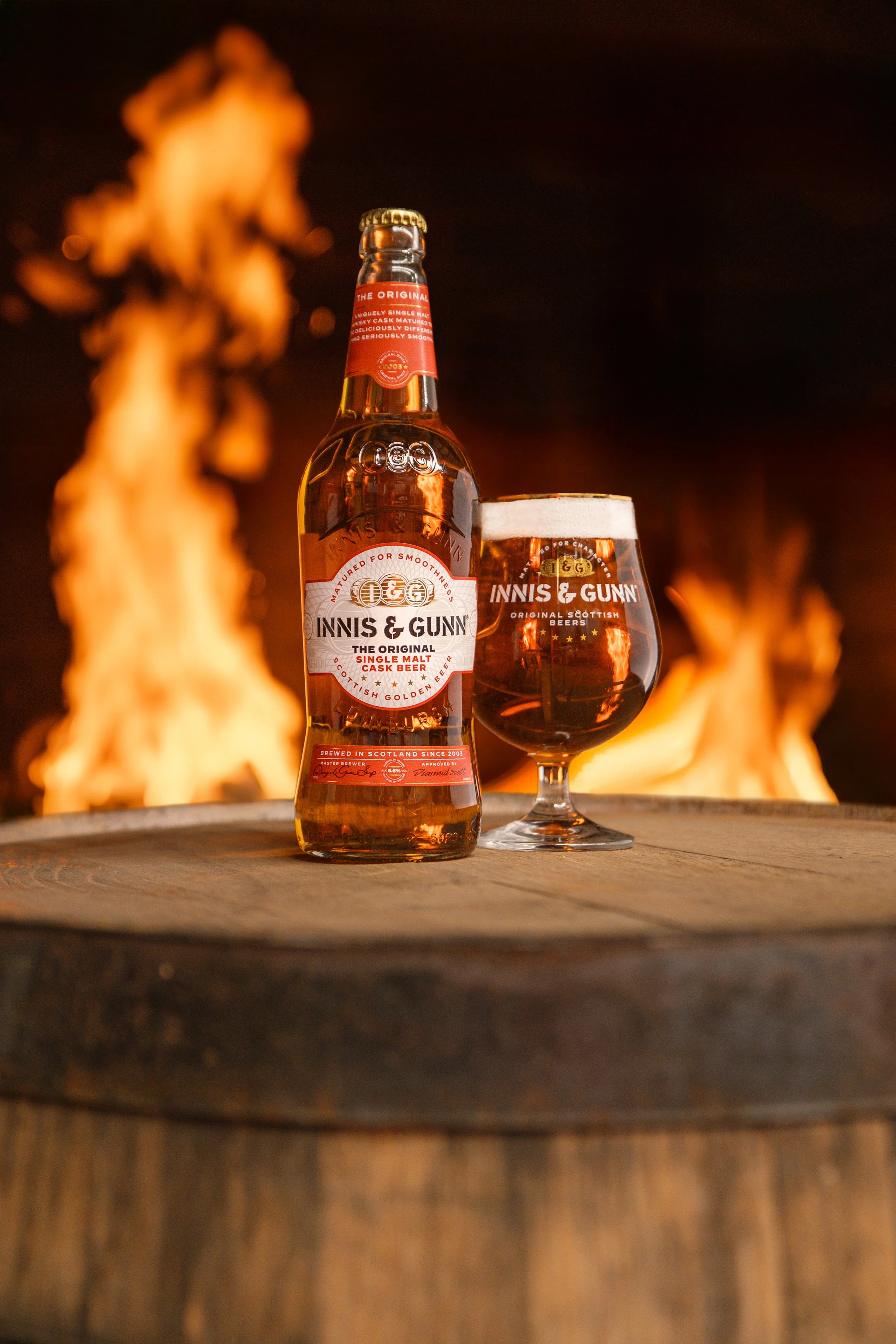 A bottle of Innis & Gunn original single malt cask beer and a glass of beer on a wooden surface with flames in the background.