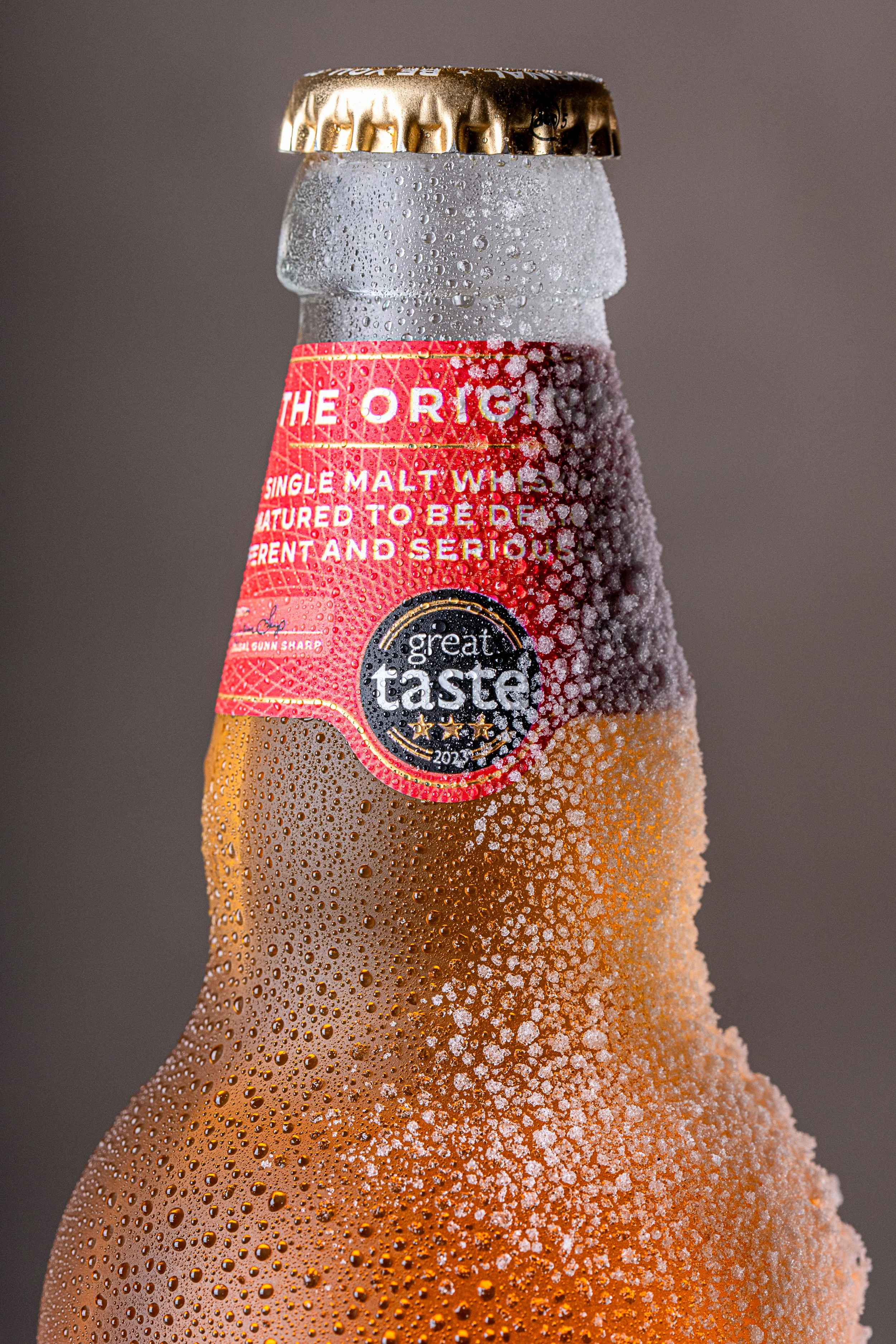 A close-up of a cold glass beer bottle covered in condensation, with a red label and a gold bottle cap.