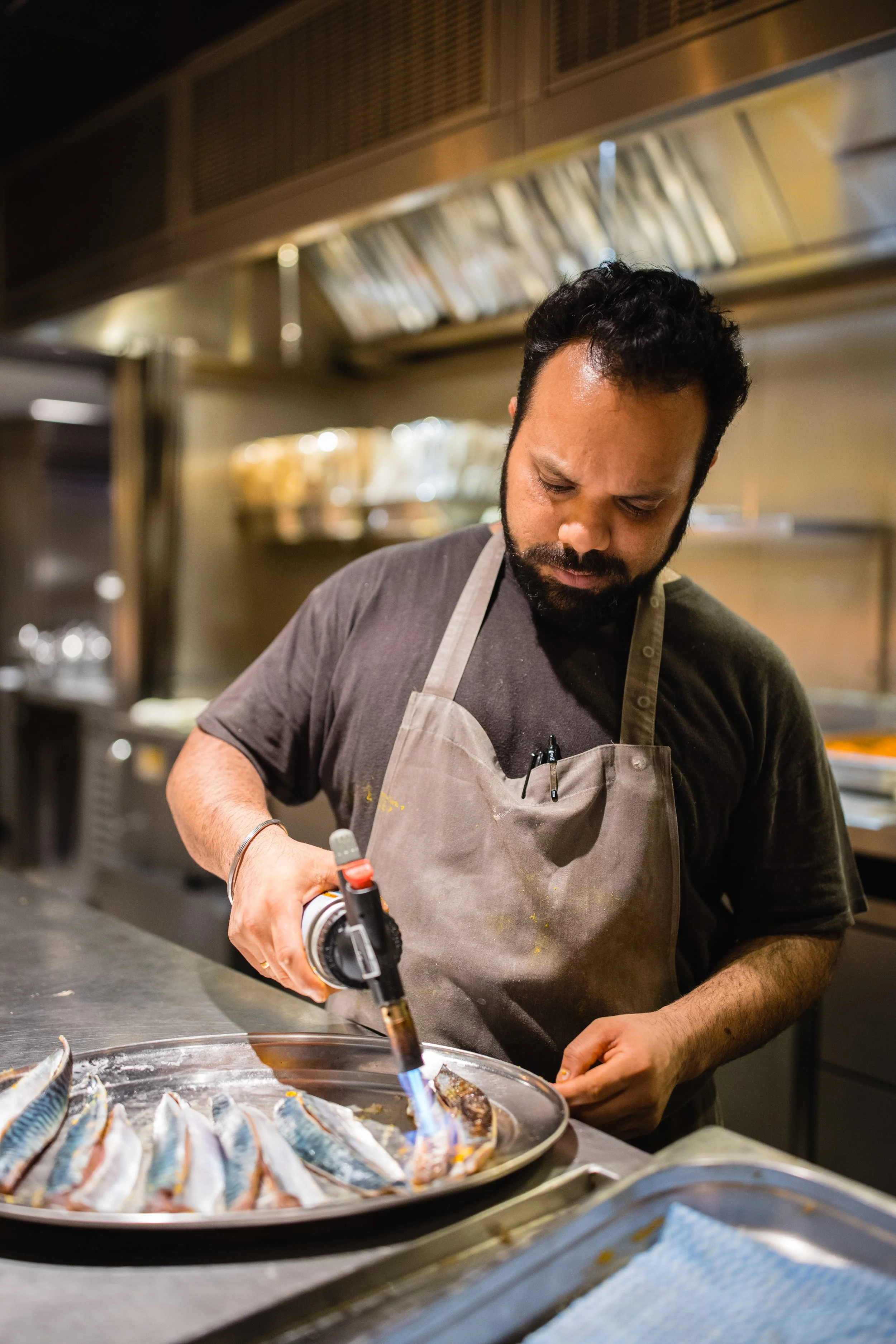 A man in a black shirt and apron is using a blowtorch to cook fish on a metal tray in a kitchen.