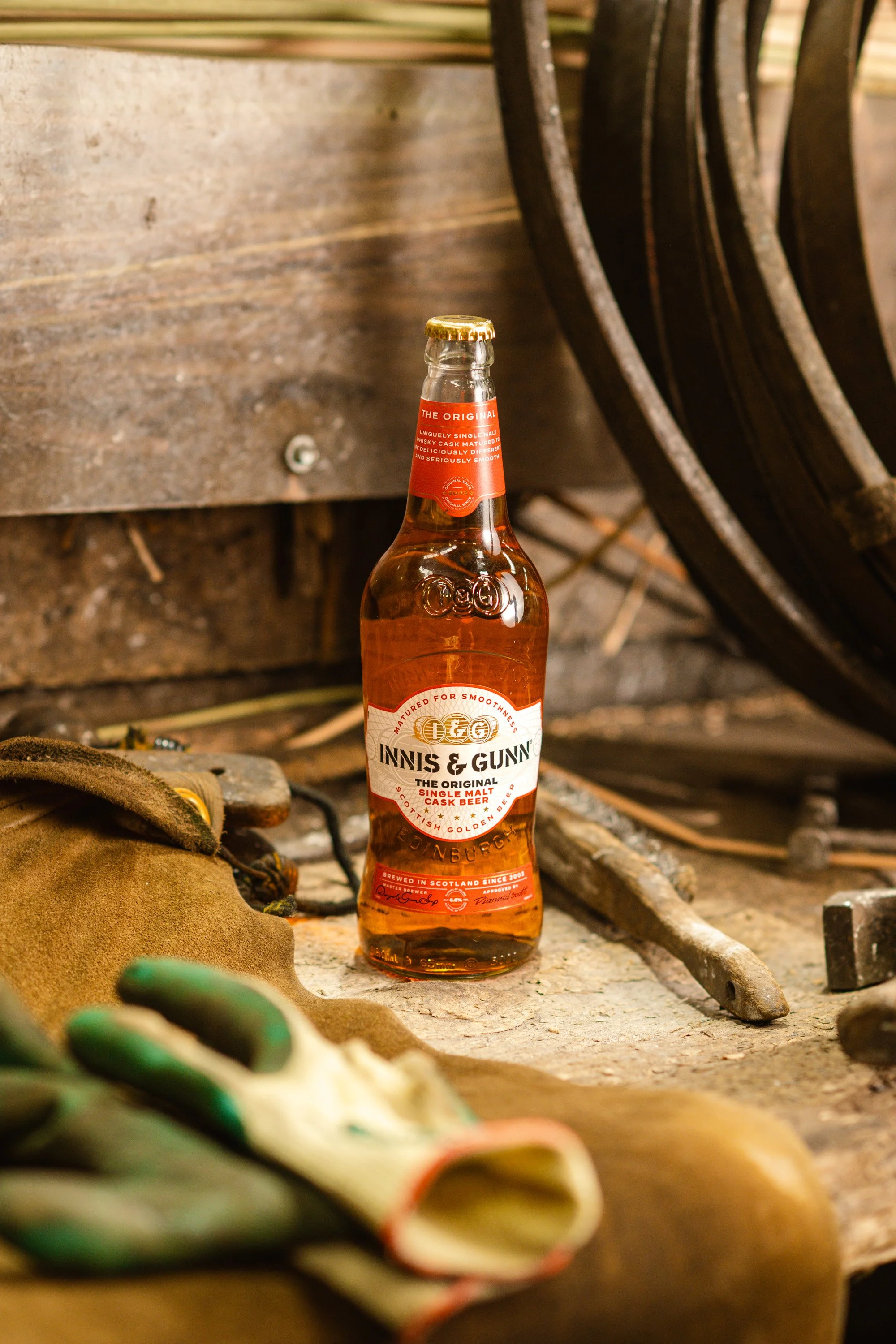 A bottle of Innis & Gunn The Original single malt cask beer is placed on a rustic wooden surface among tools and work gloves, with a blurry background of woodworking or workshop items.