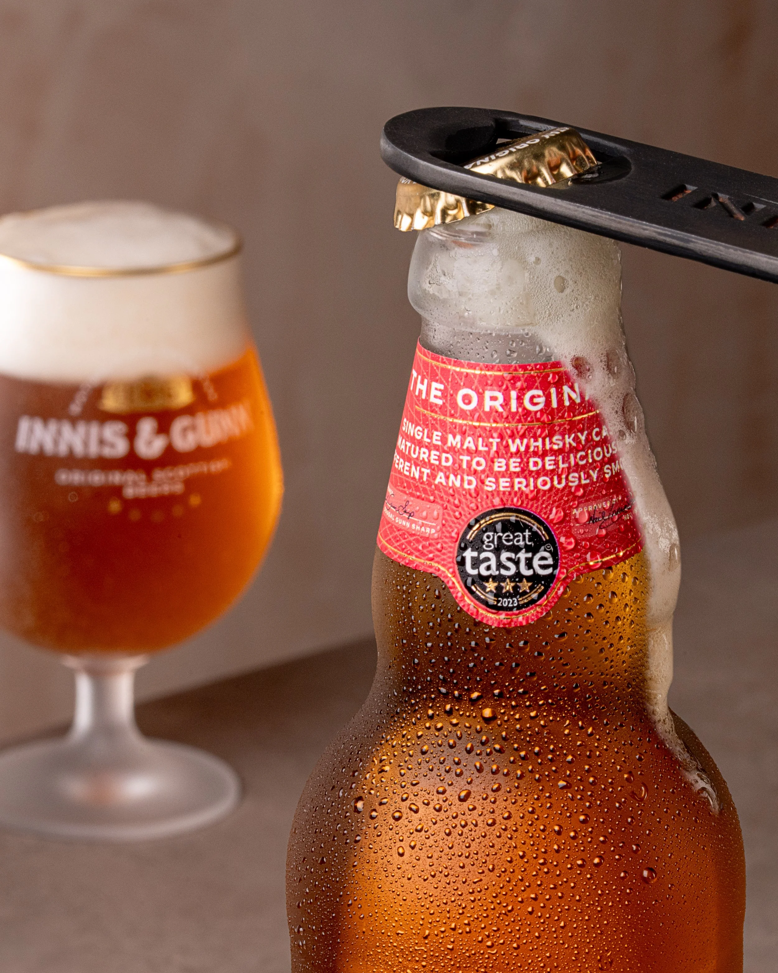 A cold, condensation-covered beer bottle with a red label and a bottle opener on top, alongside a blurred glass of beer in the background.