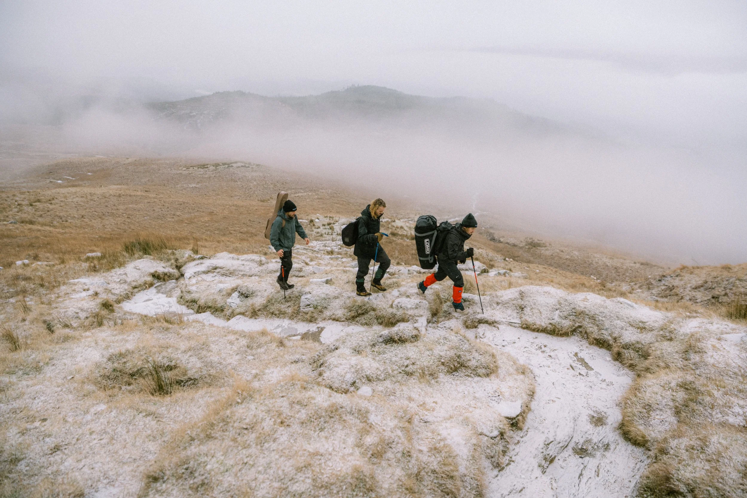 Three hikers with backpacks and trekking poles walking along a rugged mountain trail on a foggy day.