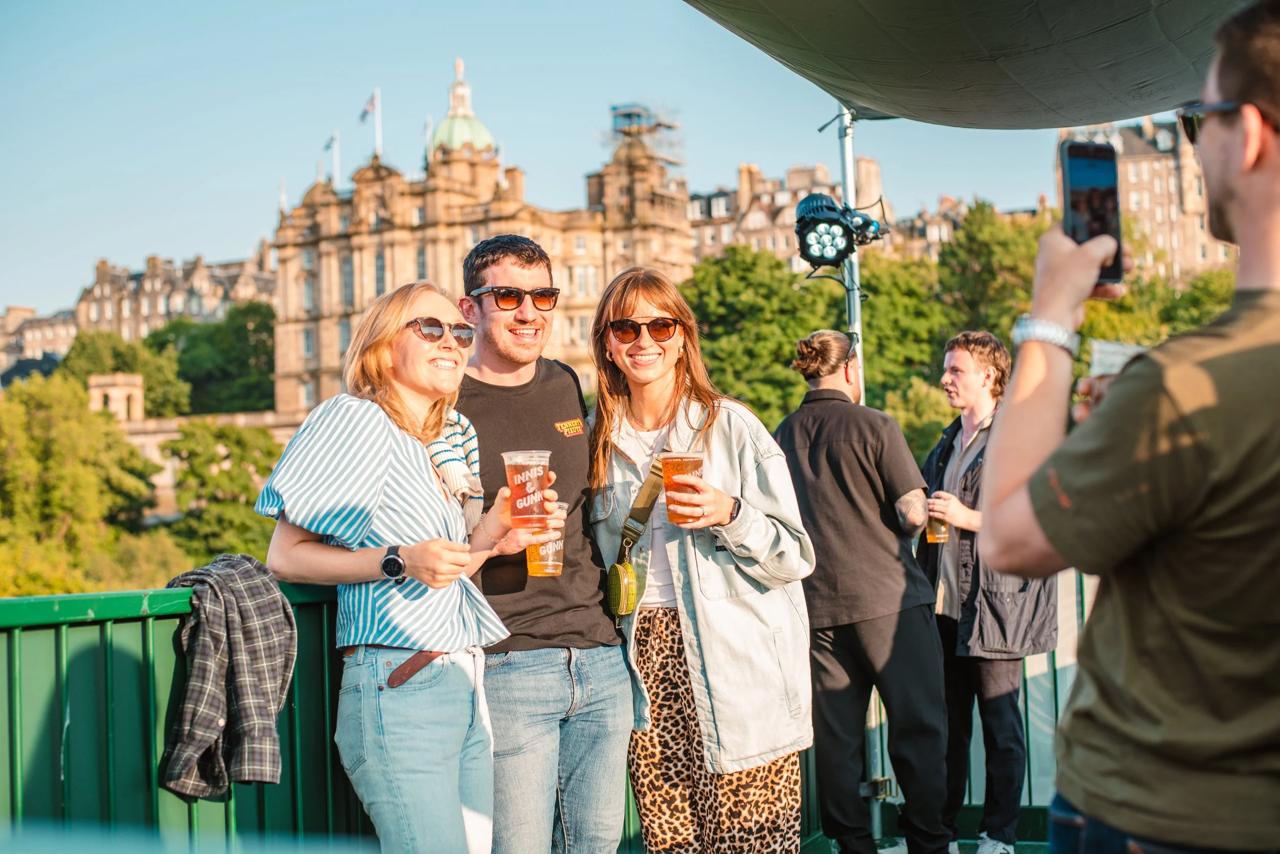 Three friends with drinks smiling for a photo on a boat deck with a historic building and green trees in the background during daytime.