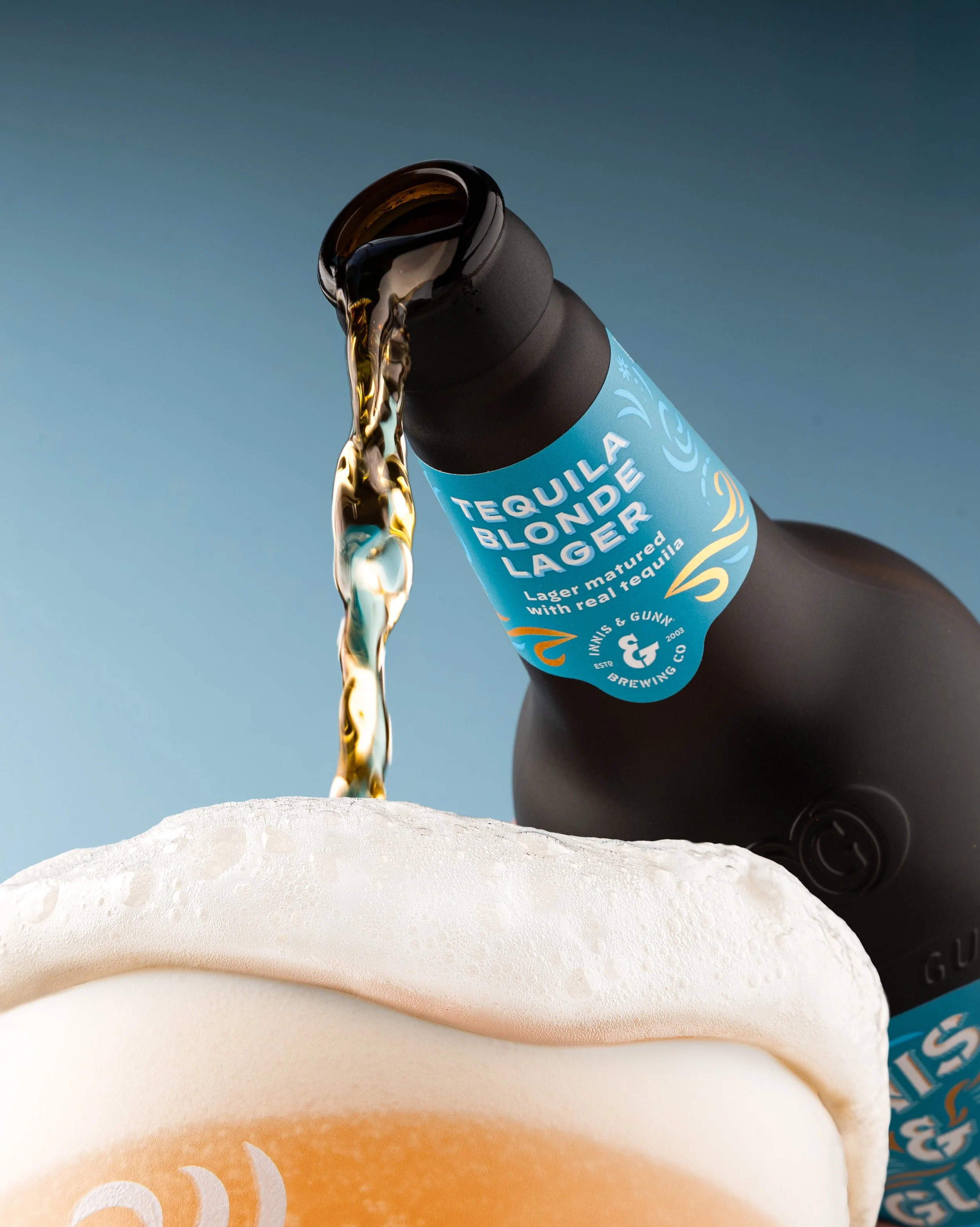 A black bottle of Tequila Blonde Lager is pouring beer into a glass, with foam at the top, against a blue background.