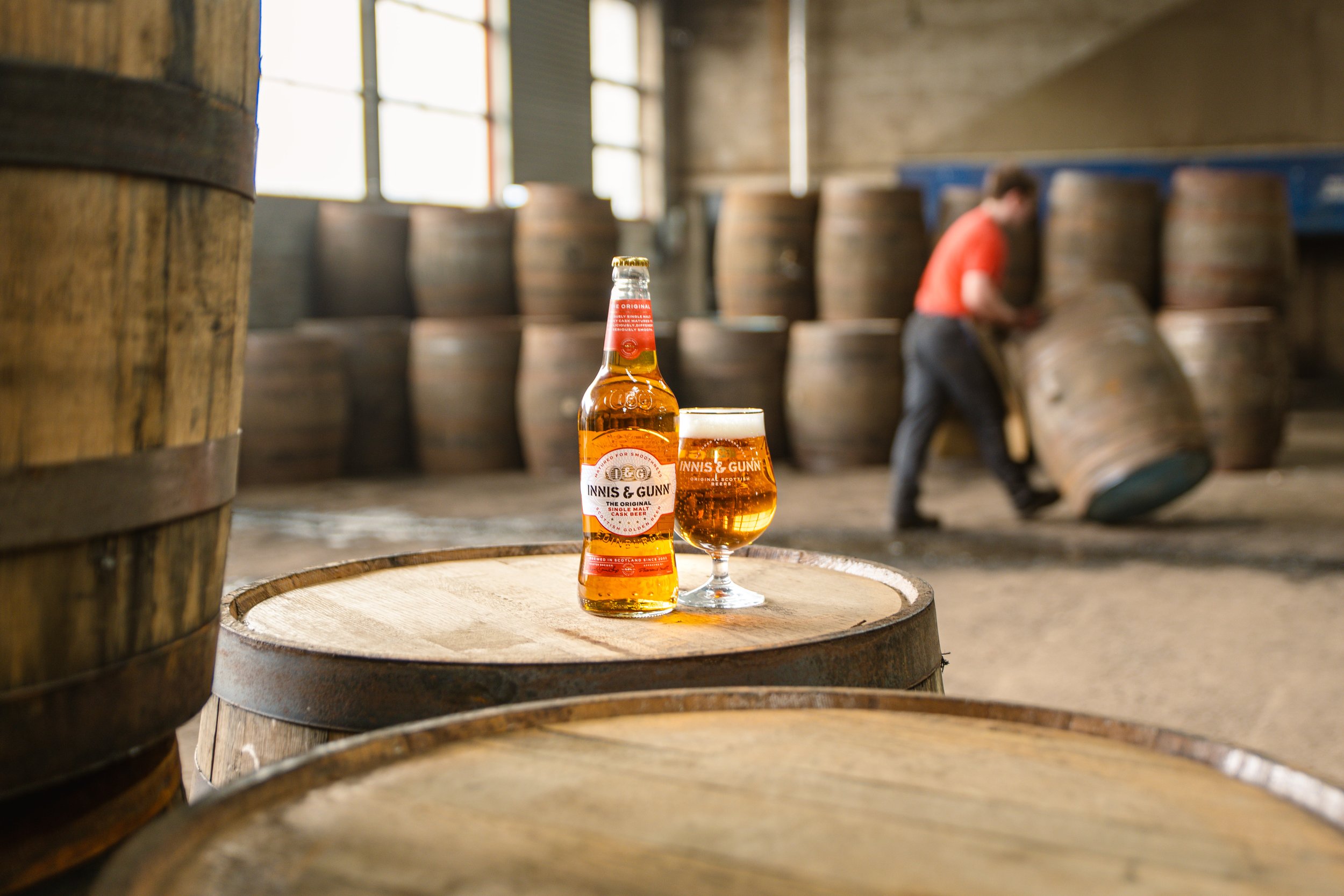 A bottle of Inniskillin & Gunn beer and a glass filled with beer on a wooden barrel in a whiskey distillery, with a worker in the background stacking barrels.