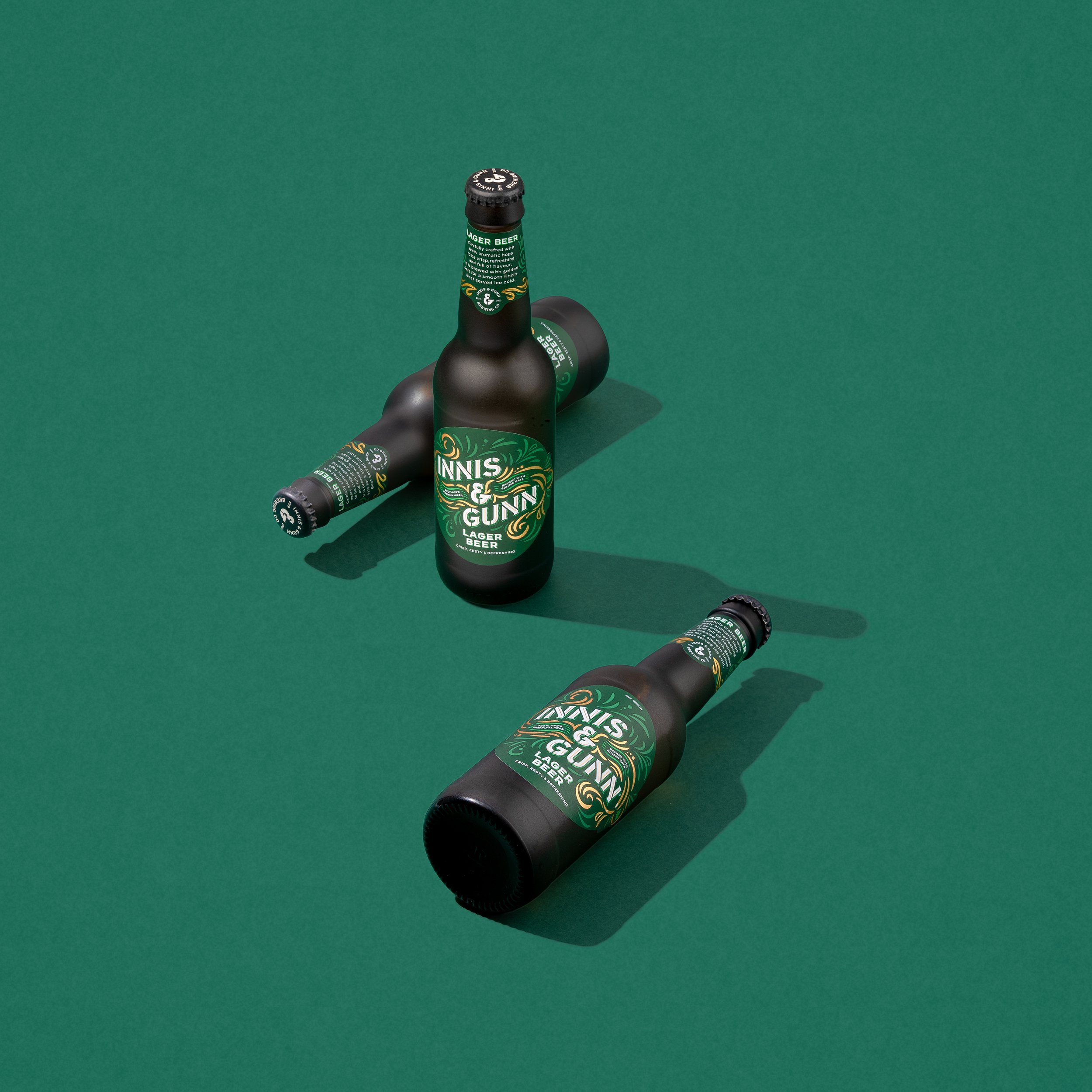 Three black bottles of Innis & Gunn lager beer with green and gold labels placed on a green surface, casting shadows.
