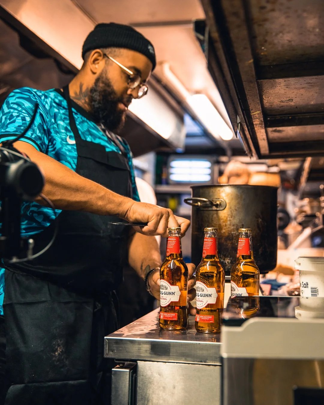 A man with a beard, glasses, and a beanie hat opening a bottle of beer in a kitchen. Three bottles of beer are lined up on the counter in front of him. He is wearing a blue patterned shirt and a black apron.