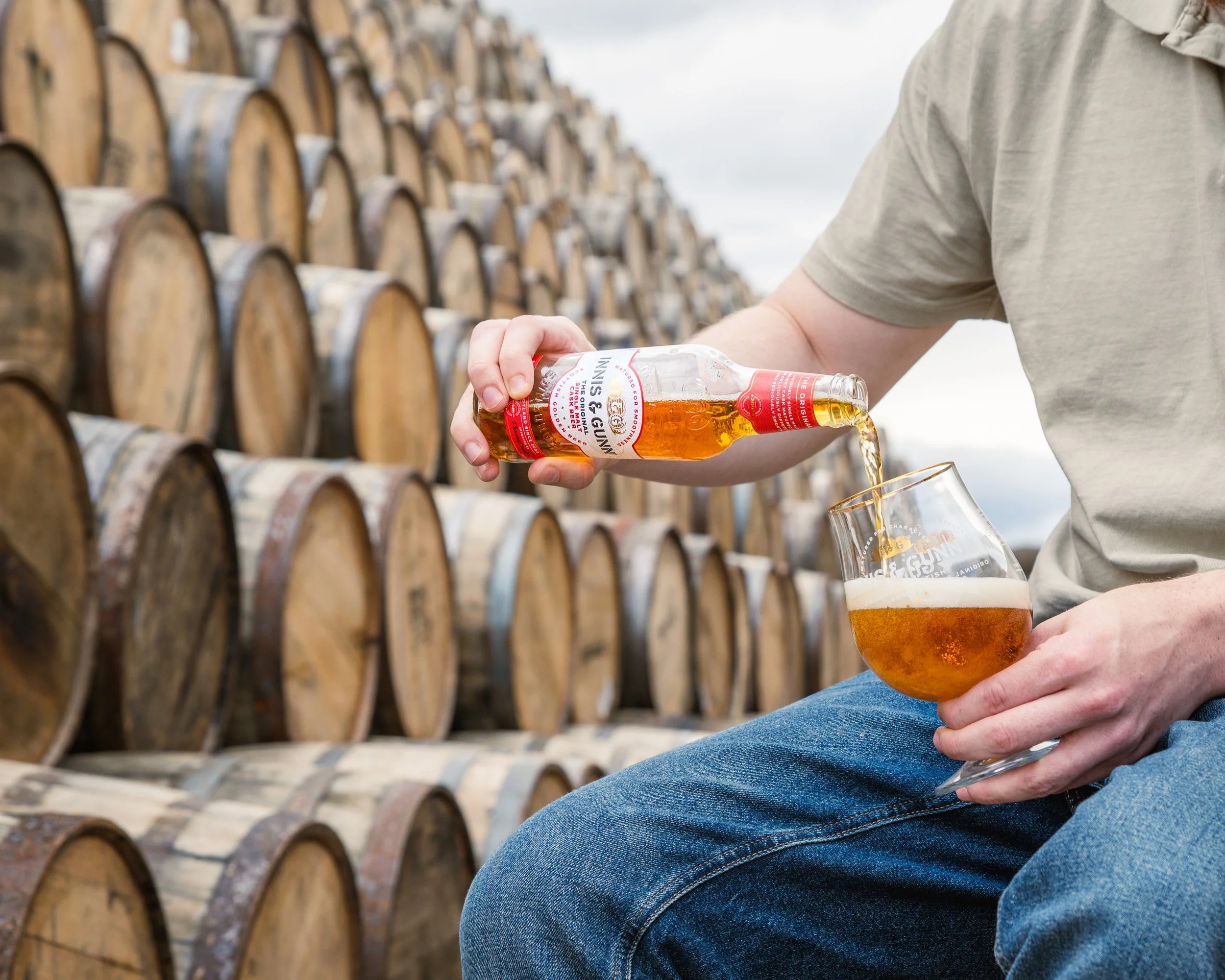 Person pouring beer from a bottle into a glass in front of stacked wooden barrels.
