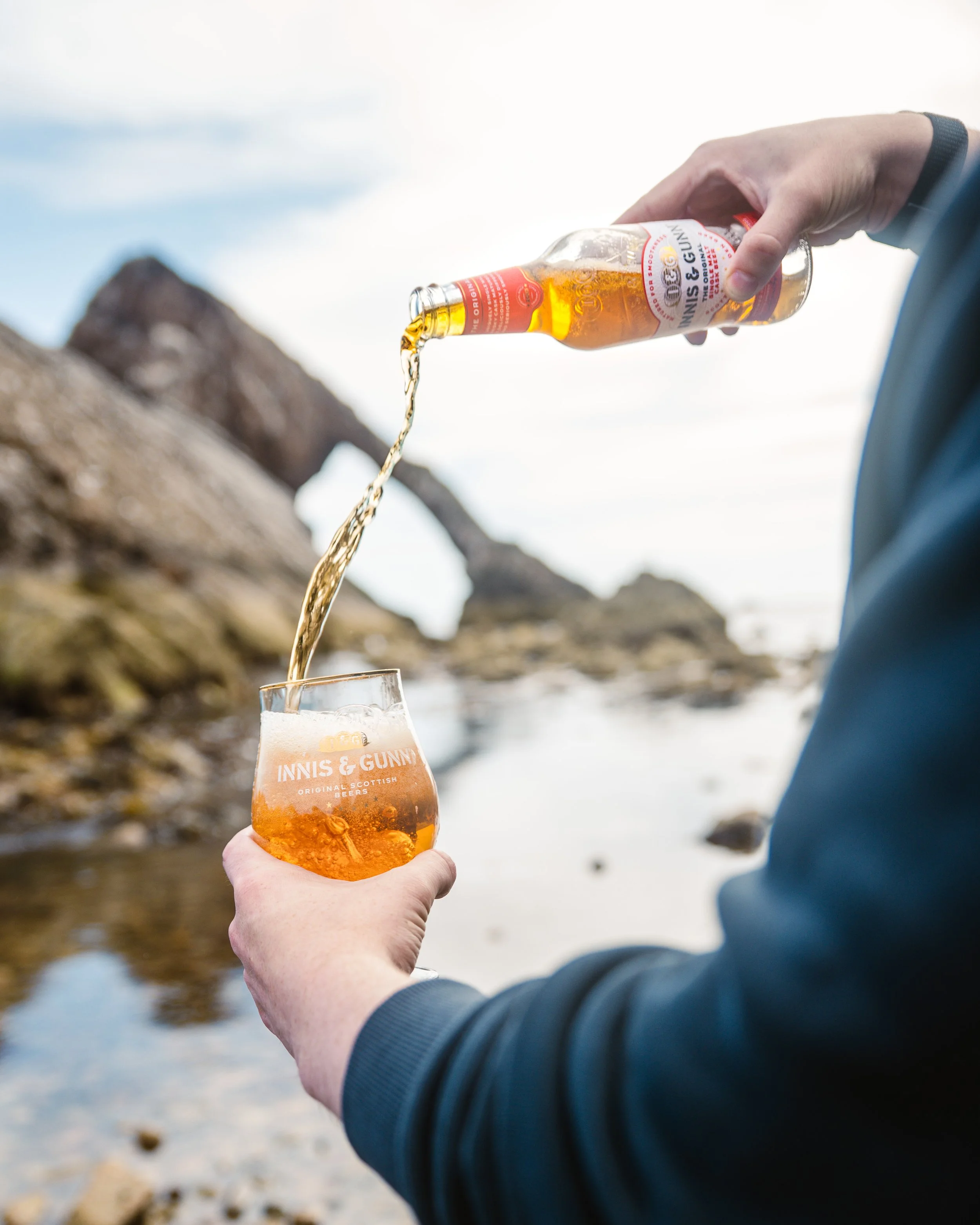 Person pouring a bottle of beer into a glass near a rocky shoreline.