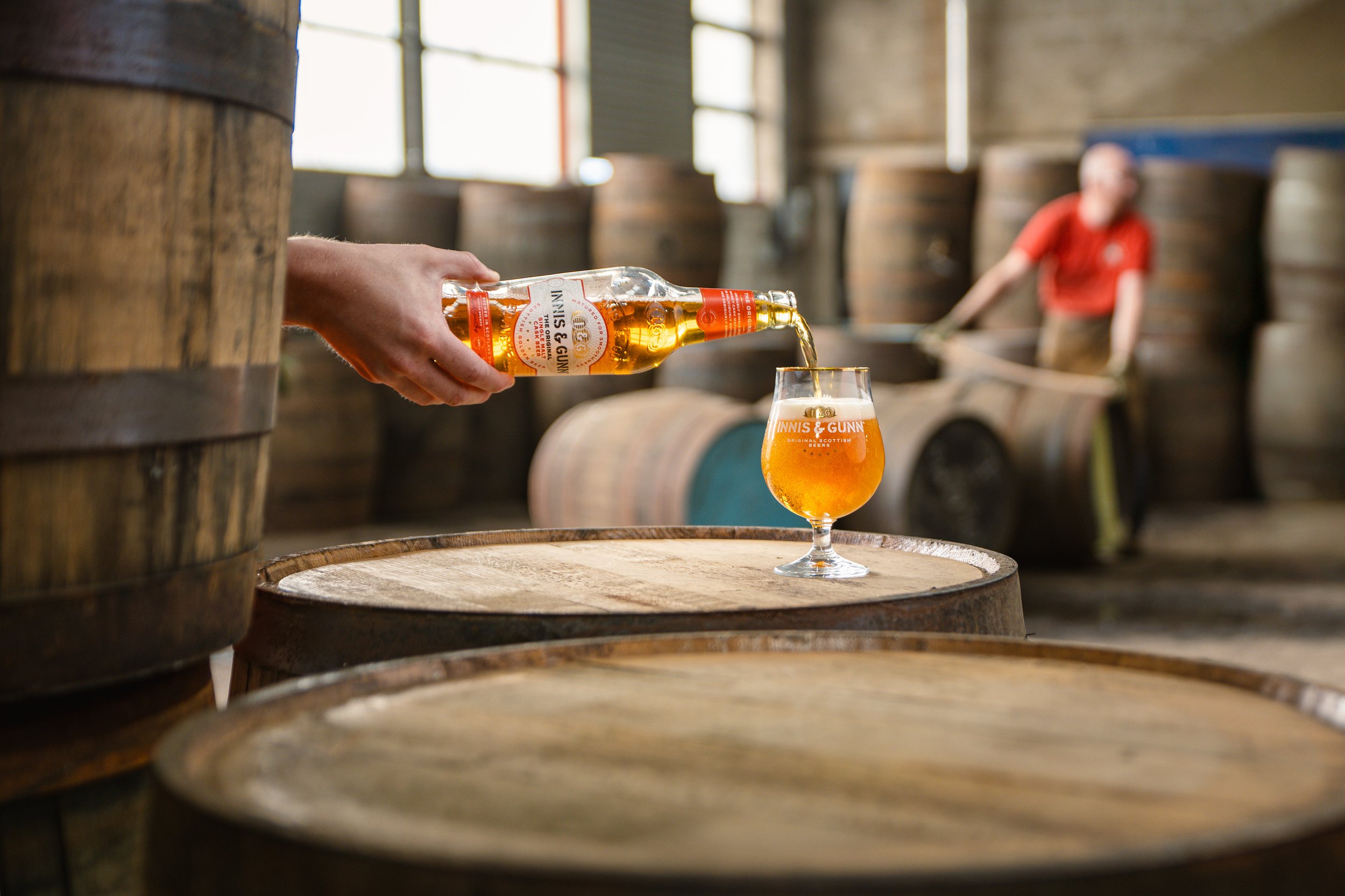 Person pouring beer into a branded glass in a distillery with wooden barrels and a worker in the background.