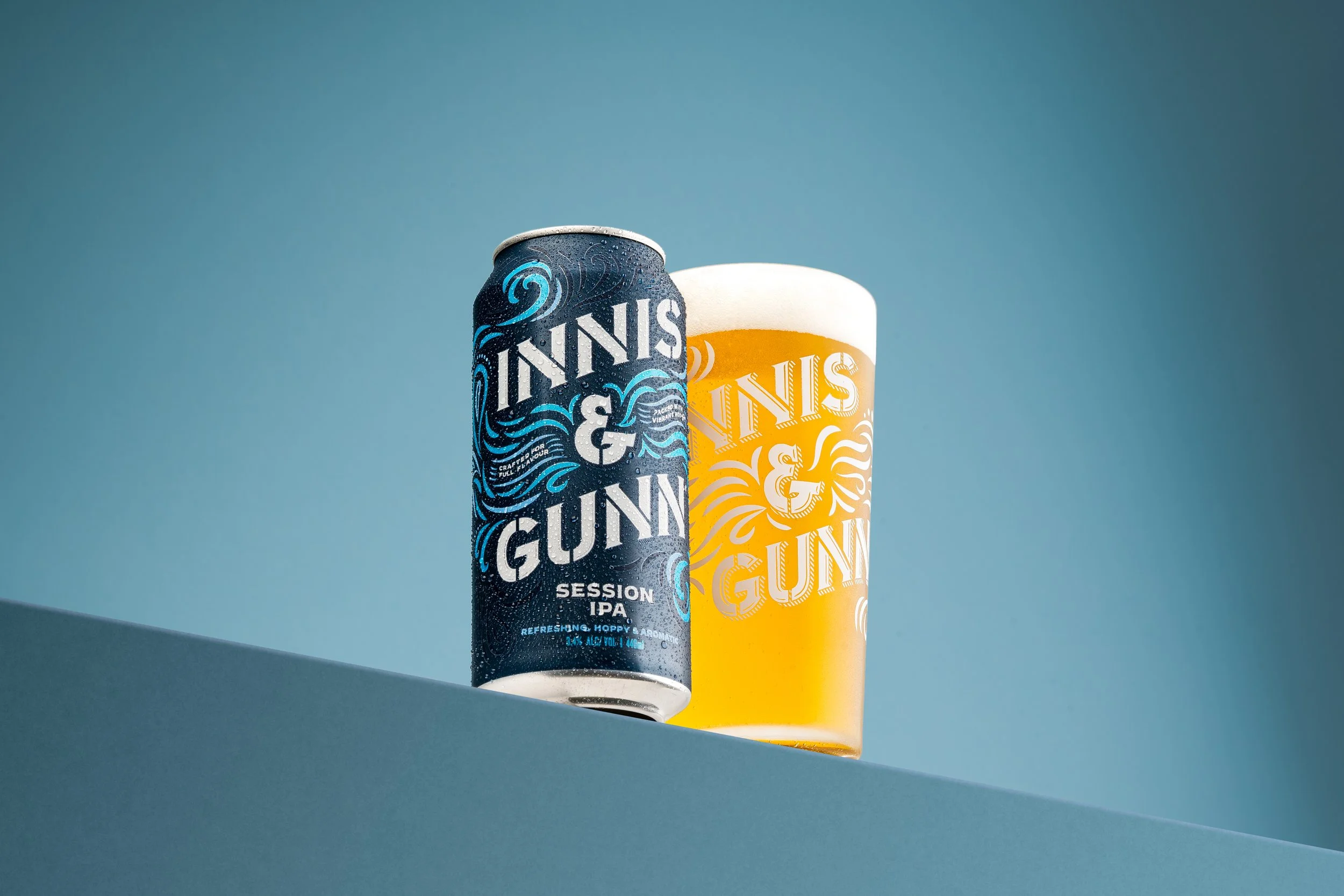 A can of Inniss & Gunni Session IPA beer and a glass of beer with foam on top, both featuring the Inniss & Gunni logo, set against a gradient blue background.