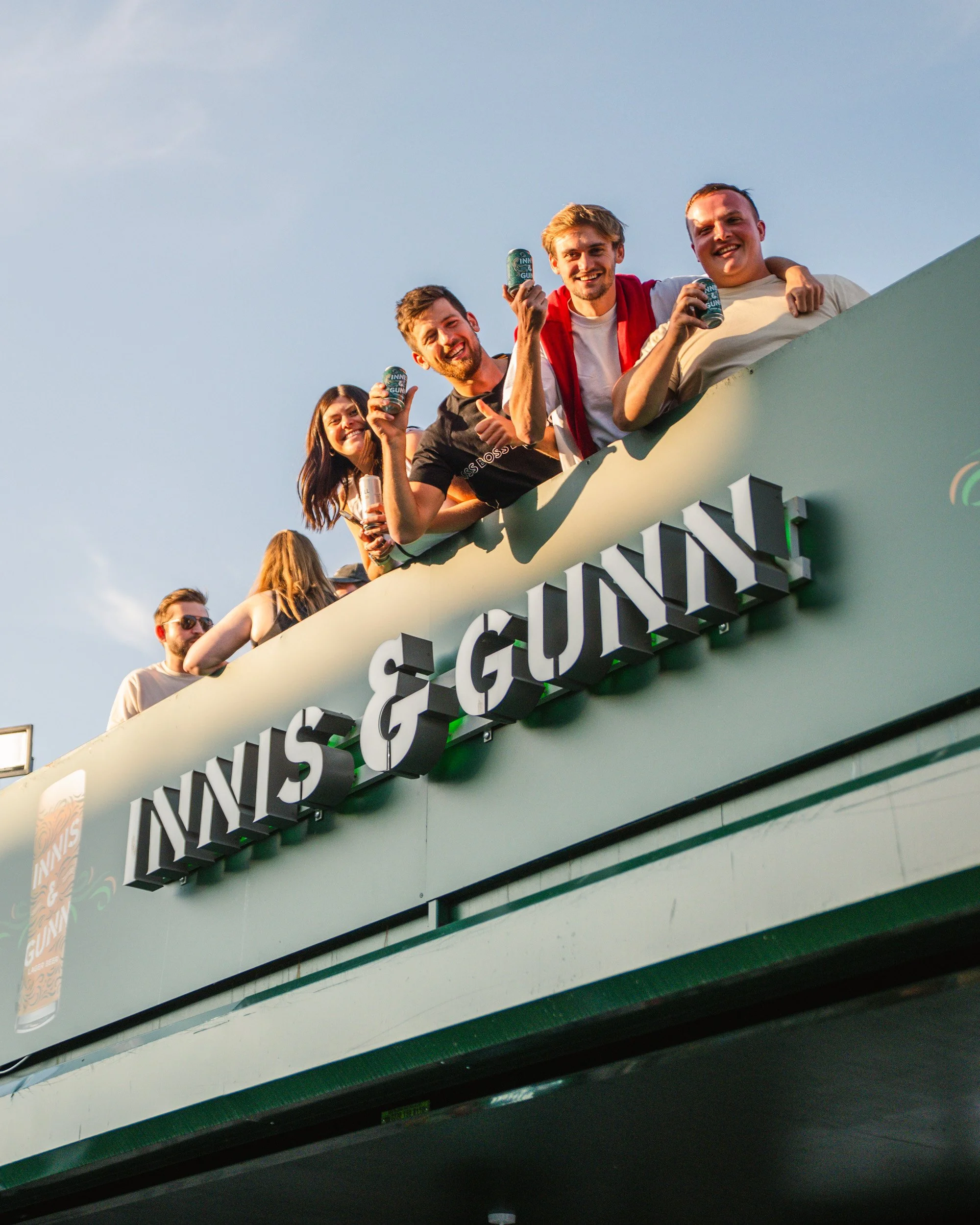 Group of friends celebrating and holding drinks on a balcony with a sign that reads 'WINS & GUNNY' during sunset.